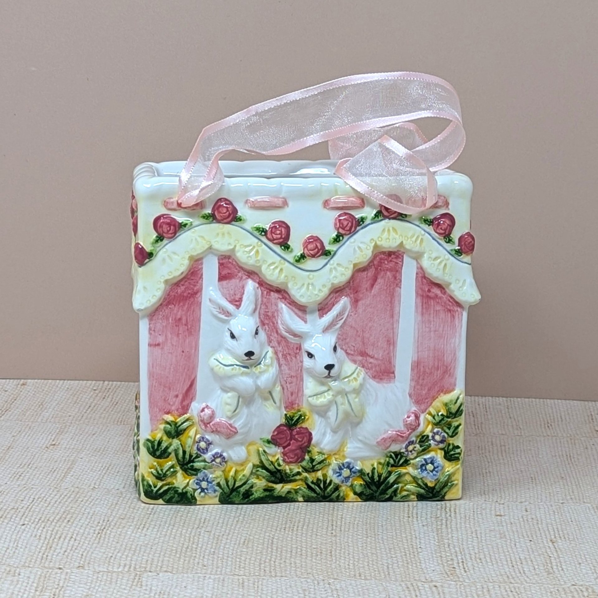 A square ceramic vase shaped like a gift bag with pink ribbon handles. The front features a high-relief scene of two white bunnies in a garden with pink roses and yellow lace detailing. Glossy vitrified texture.