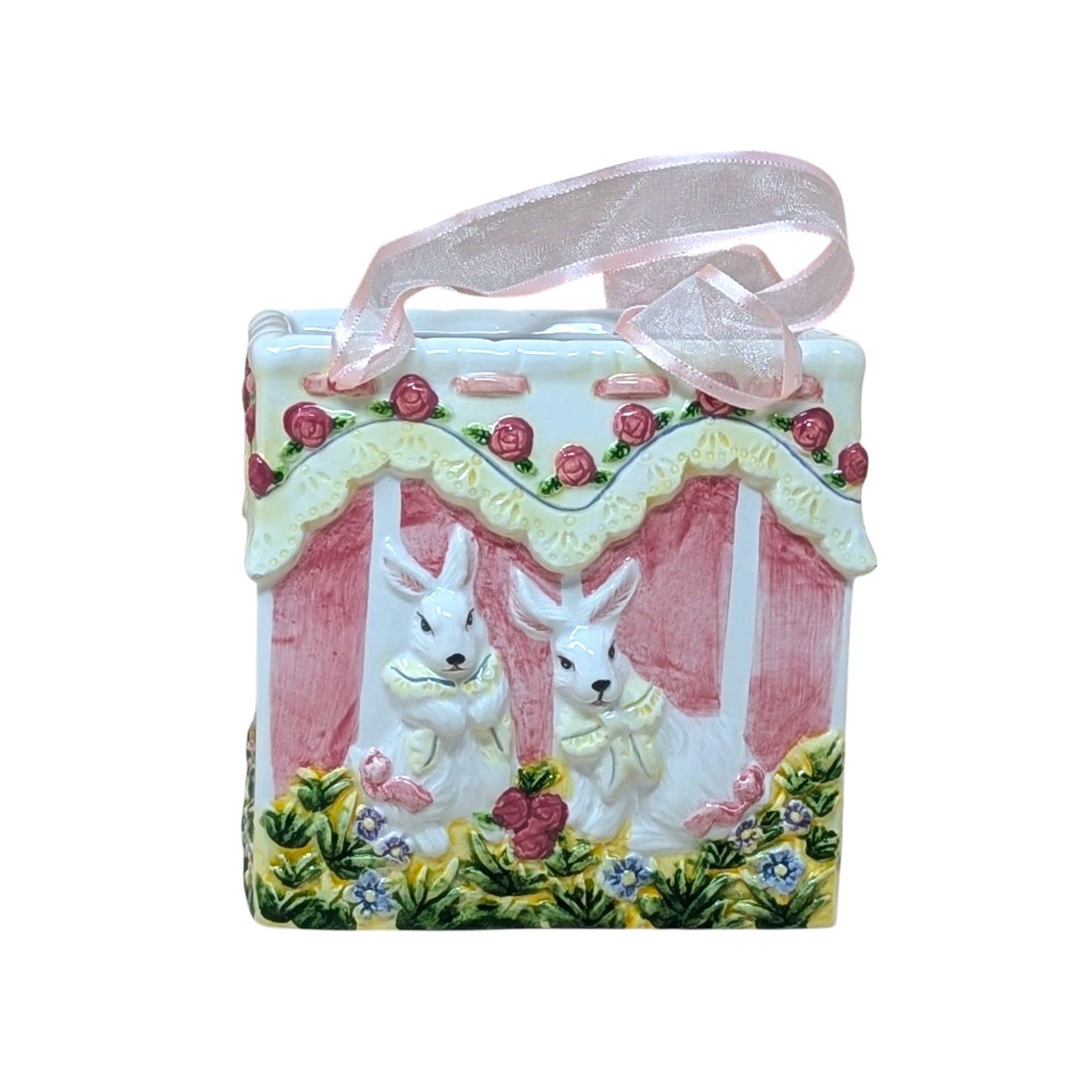 A square ceramic vase shaped like a gift bag with pink ribbon handles. The front features a high-relief scene of two white bunnies in a garden with pink roses and yellow lace detailing. Glossy vitrified texture.