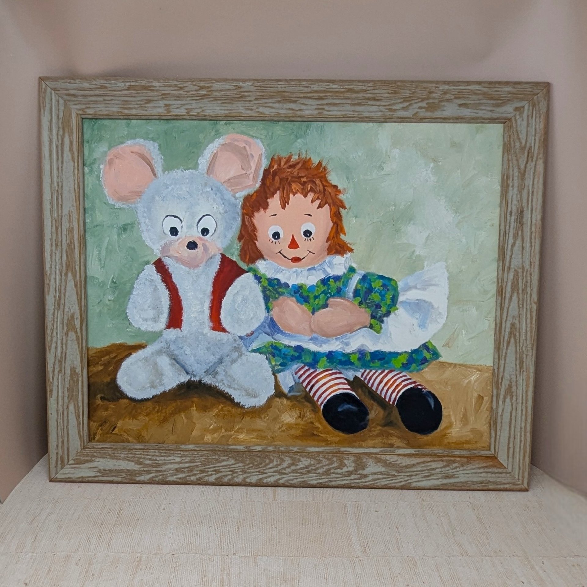 A large horizontal painting on canvas board featuring Raggedy Ann sitting next to a white teddy bear in a red vest. Impasto brushstrokes in shades of green and brown. Housed in a light wood grain frame.