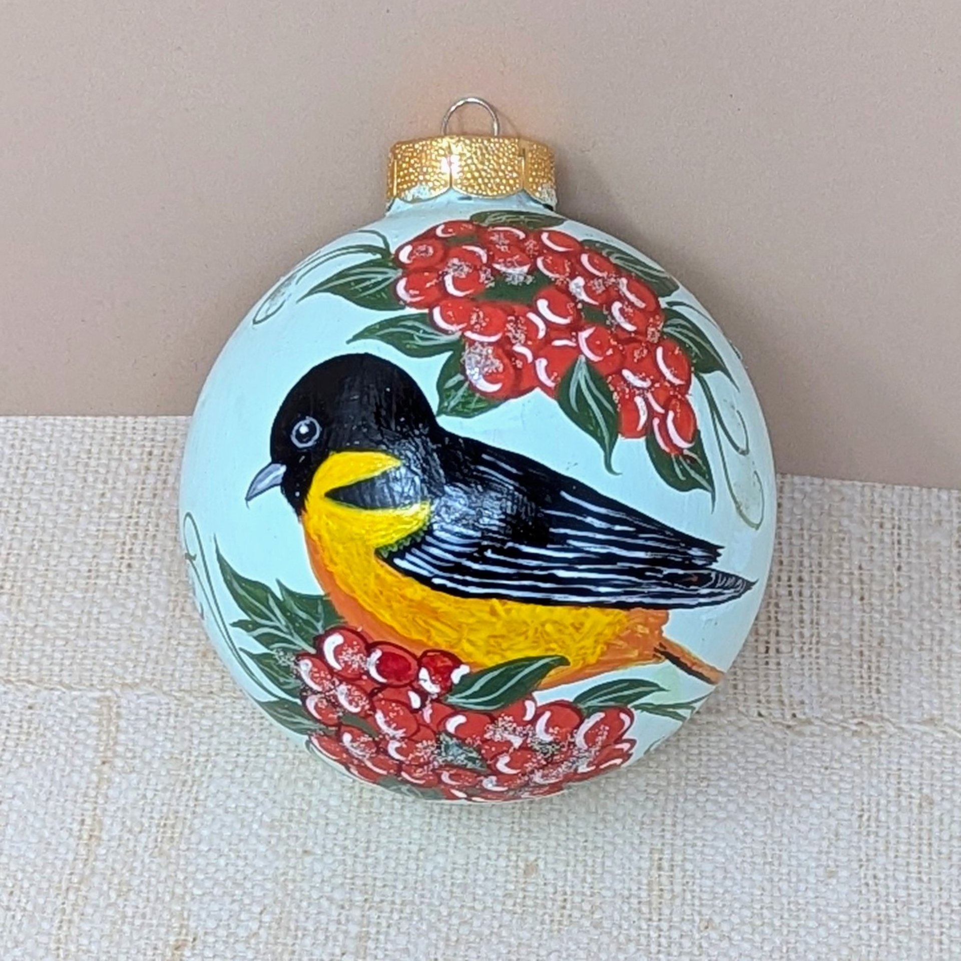 A spherical glass ornament featuring a hand-painted Baltimore Oriole bird with a black head and vibrant orange breast. Surrounded by clusters of bright red berries and green leaves. Topped with a classic gold metal cap.