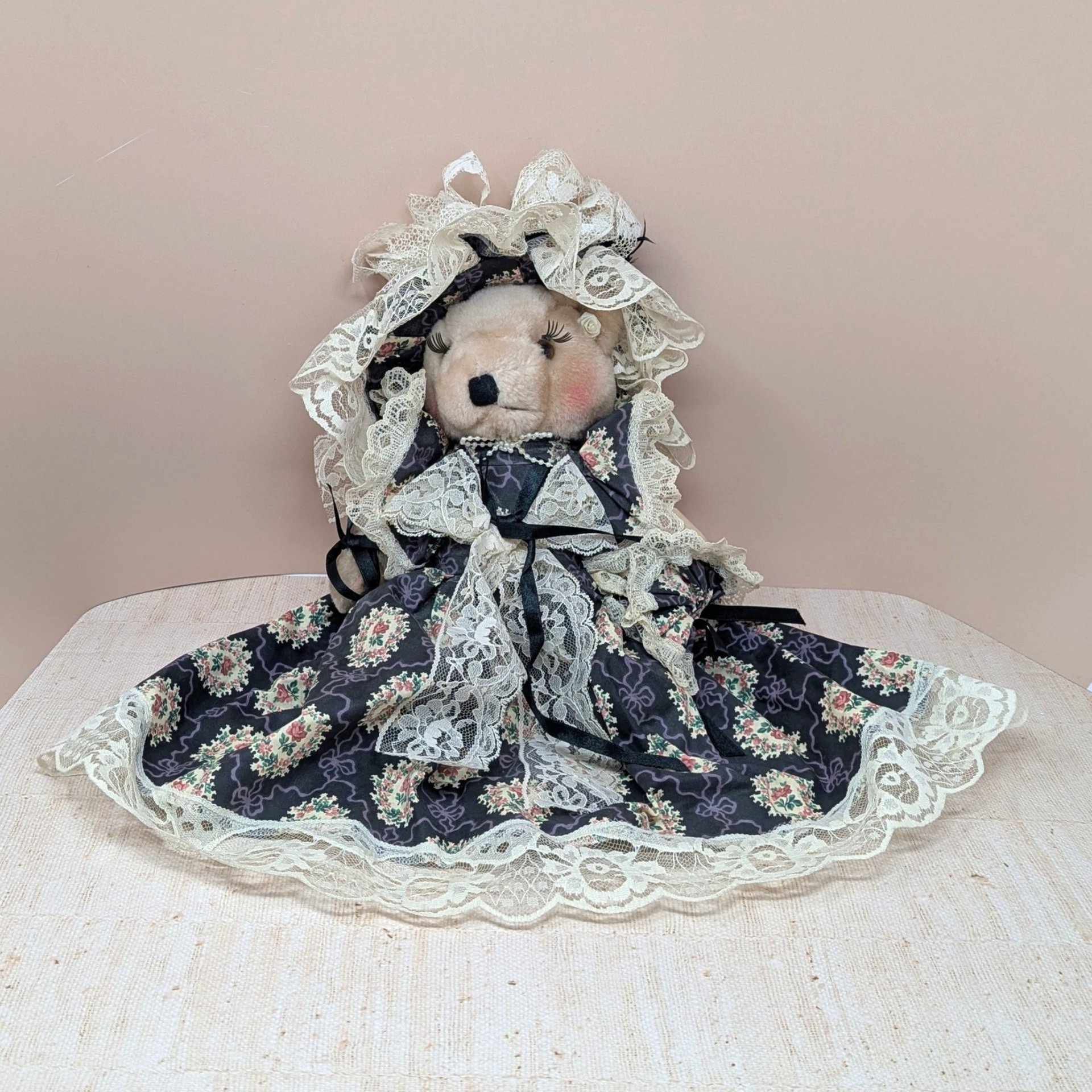 A vertical 12-inch handcrafted teddy bear with tan fur and movable limbs. Wearing an elaborate Victorian-style dark floral dress with voluminous cream lace trim and a matching oversized lace bonnet.