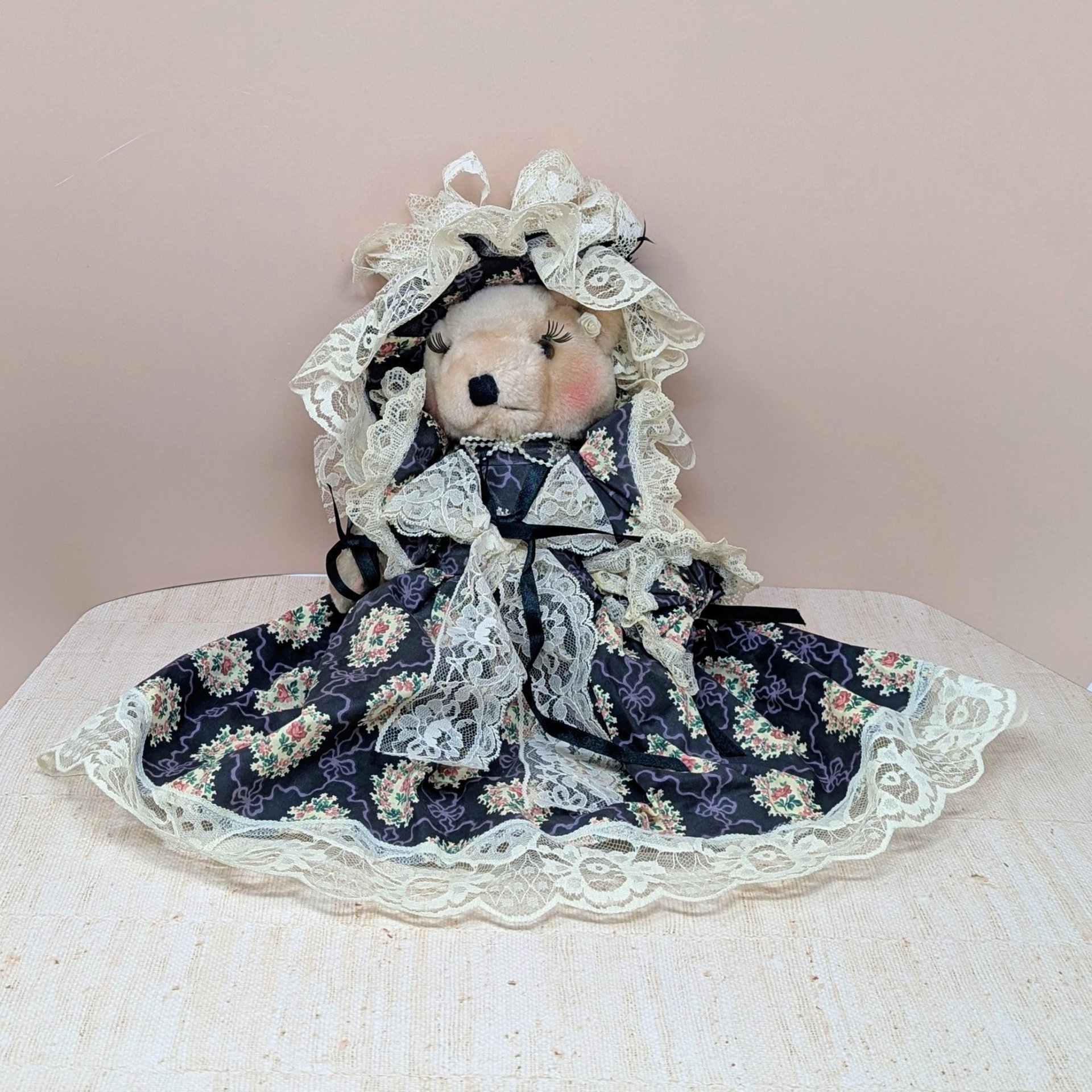 A vertical 12-inch handcrafted teddy bear with tan fur and movable limbs. Wearing an elaborate Victorian-style dark floral dress with voluminous cream lace trim and a matching oversized lace bonnet.