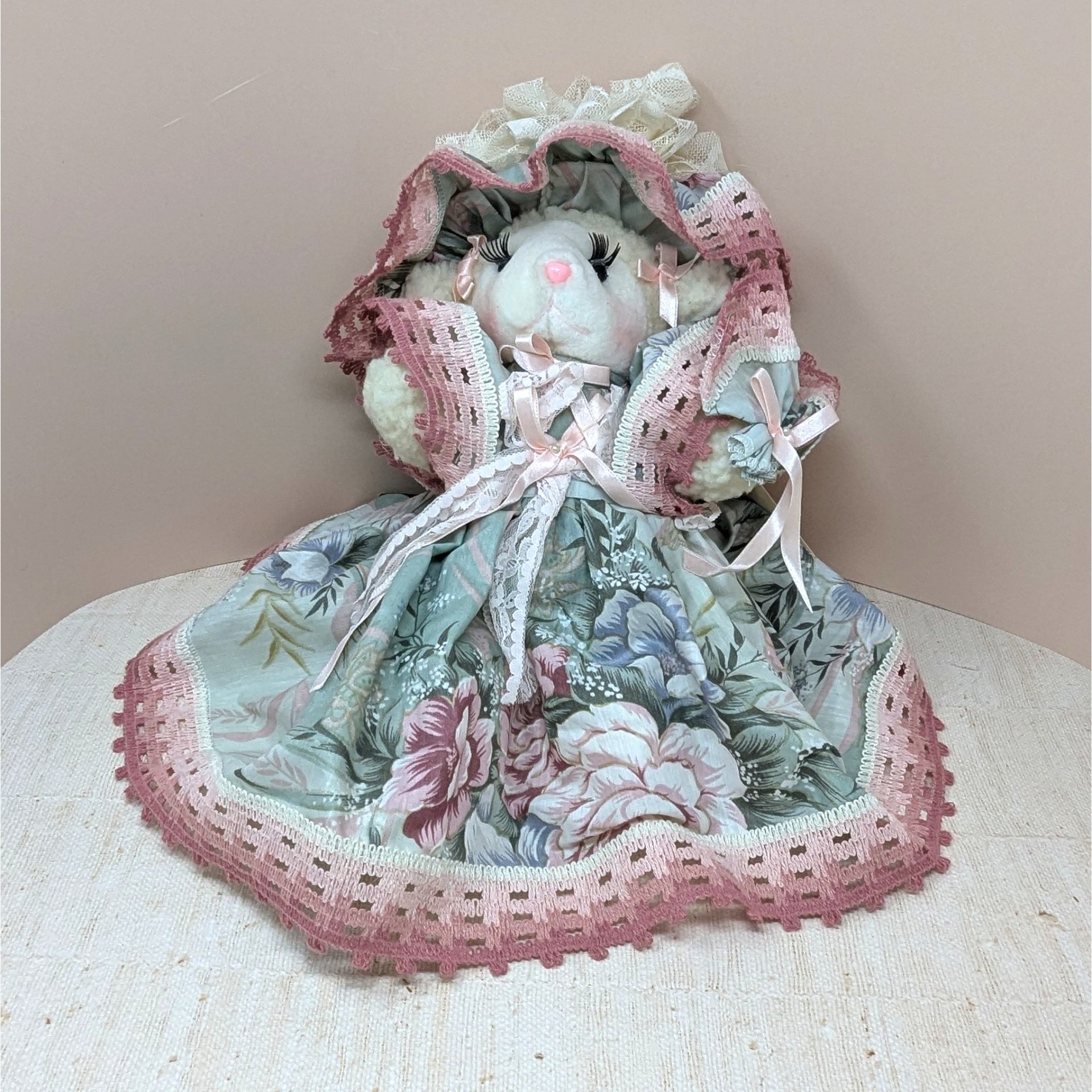 A seated plush lamb in a wide, floral-patterned sage green and rose pink dress. The lamb features long eyelashes, a pink nose, and an oversized matching lace bonnet. The intricate pink lace trim on the hem and bonnet adds a grounded, romantic texture.