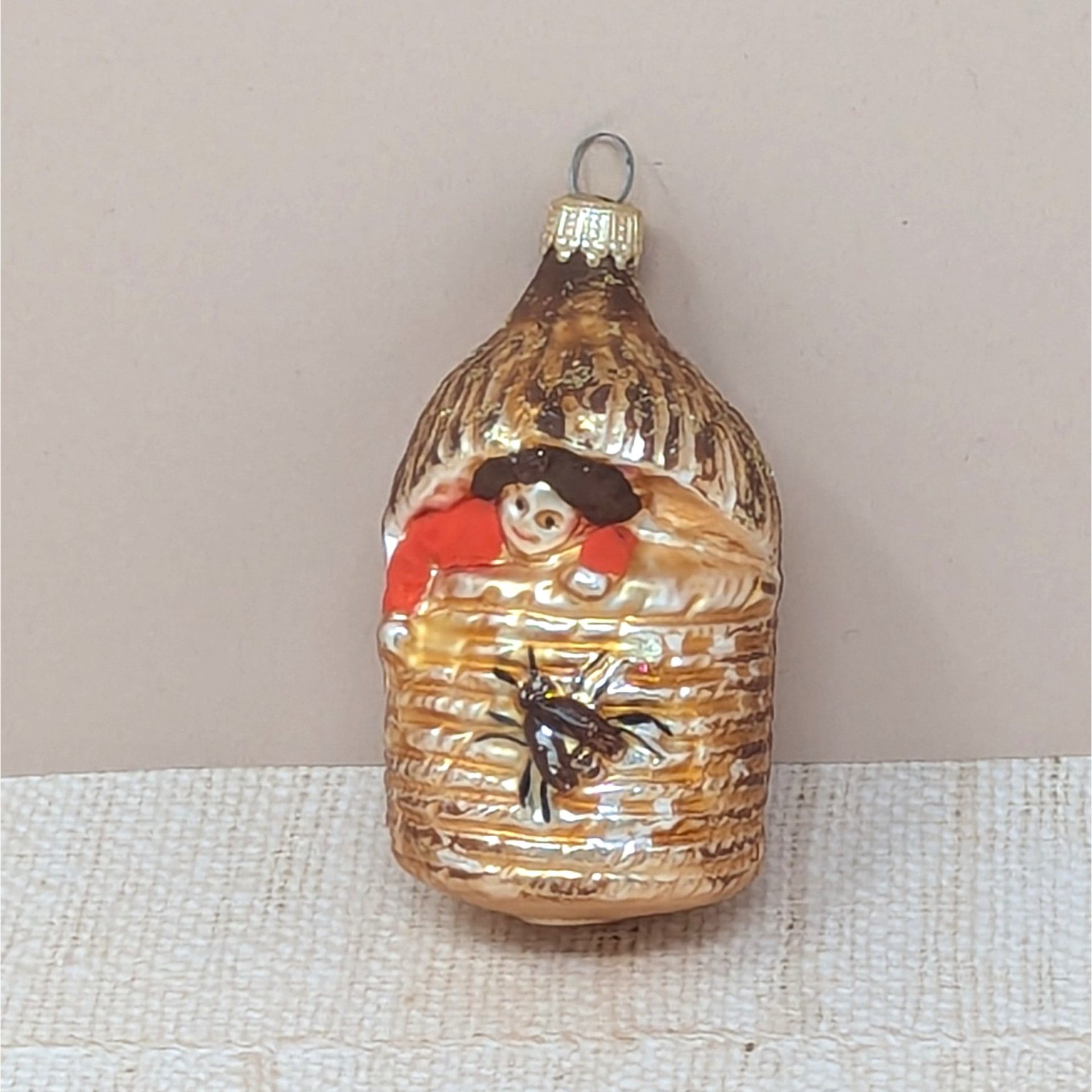 A vertical 4.5-inch vintage blown glass ornament shaped like a beehive. A hand-painted baby with a red suit peeks from the top, and a large brown bee is featured on the front. Silvered glass with gold glitter accents.