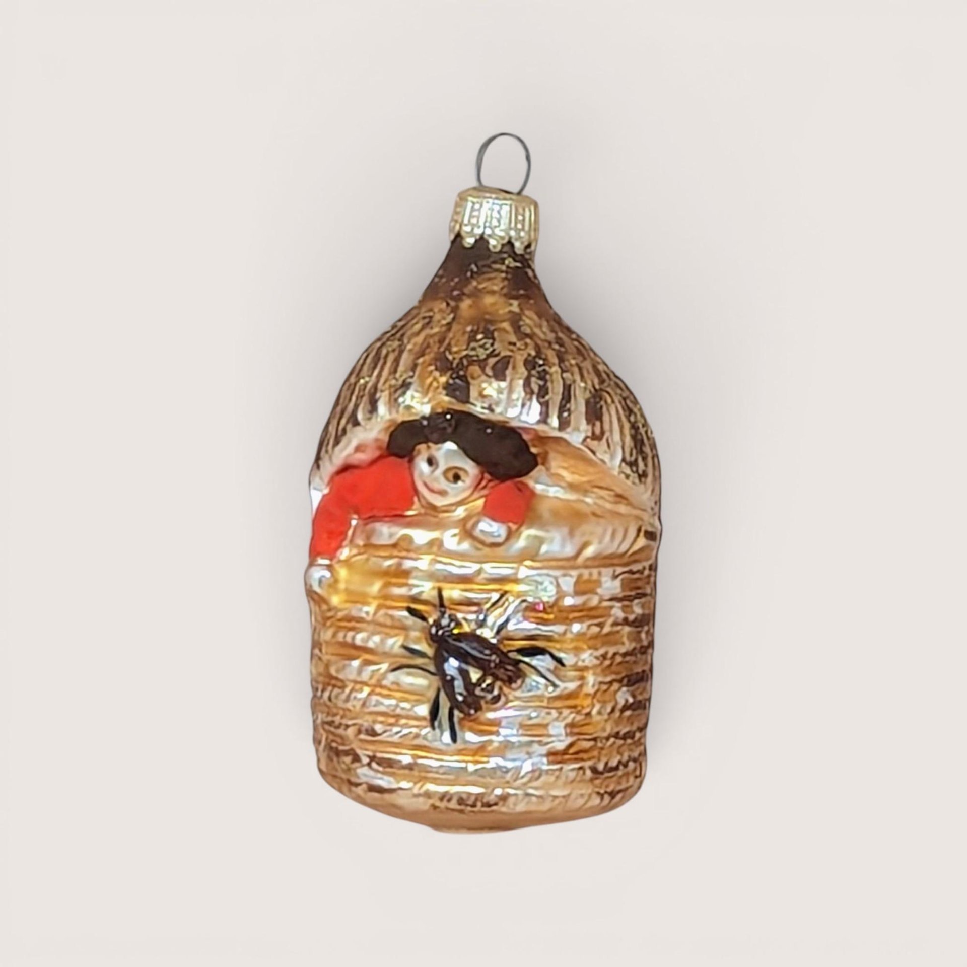 A vertical 4.5-inch vintage blown glass ornament shaped like a beehive. A hand-painted baby with a red suit peeks from the top, and a large brown bee is featured on the front. Silvered glass with gold glitter accents.