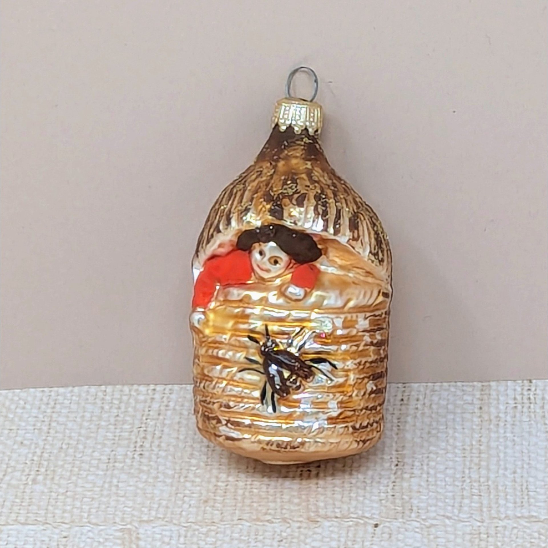 A vertical 4.5-inch vintage blown glass ornament shaped like a beehive. A hand-painted baby with a red suit peeks from the top, and a large brown bee is featured on the front. Silvered glass with gold glitter accents.