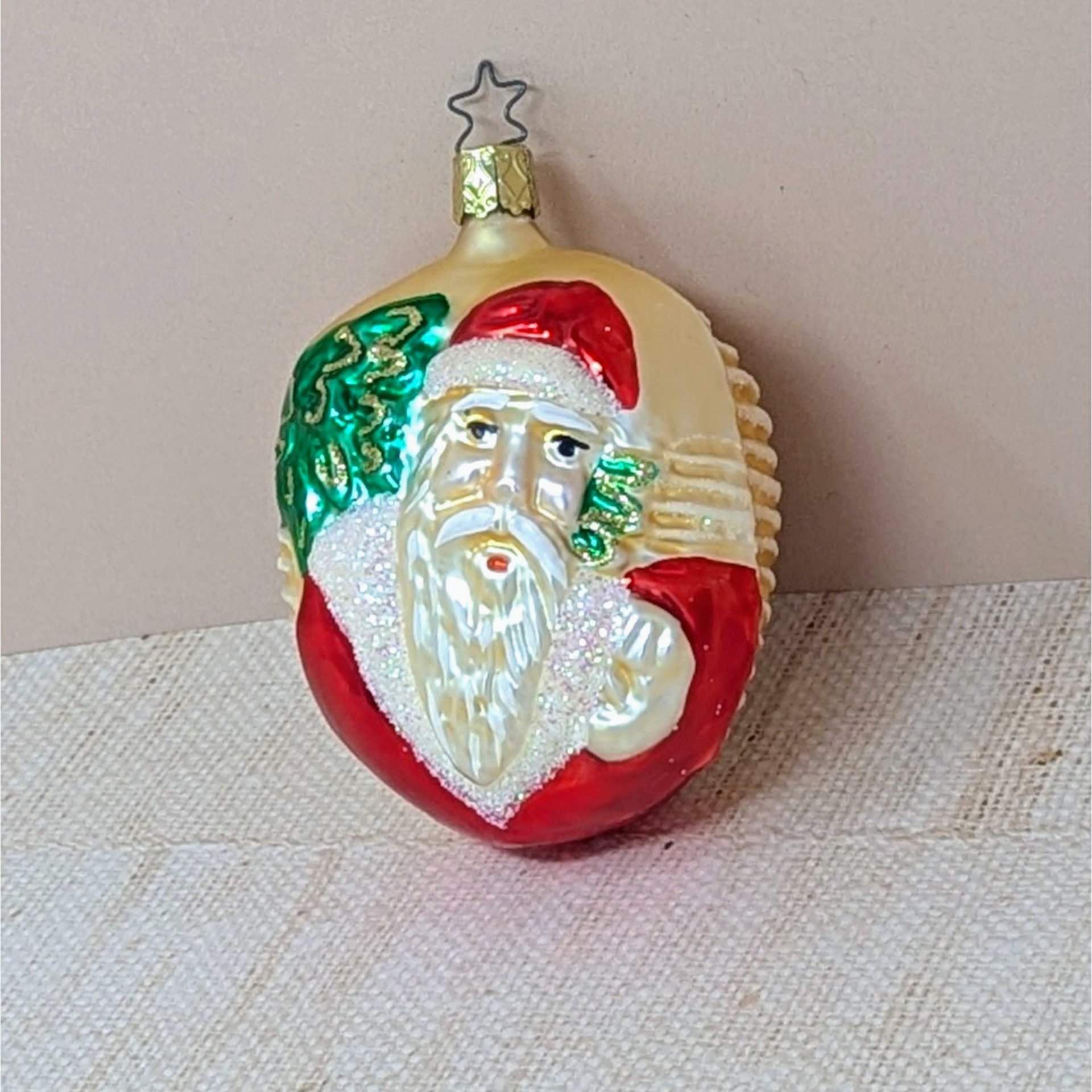 A vertical 4.5-inch blown glass ornament featuring a detailed Santa Claus face with a snowy glittered beard. He is carrying a green glittered tree. Features a signature star-shaped metal hanging loop.