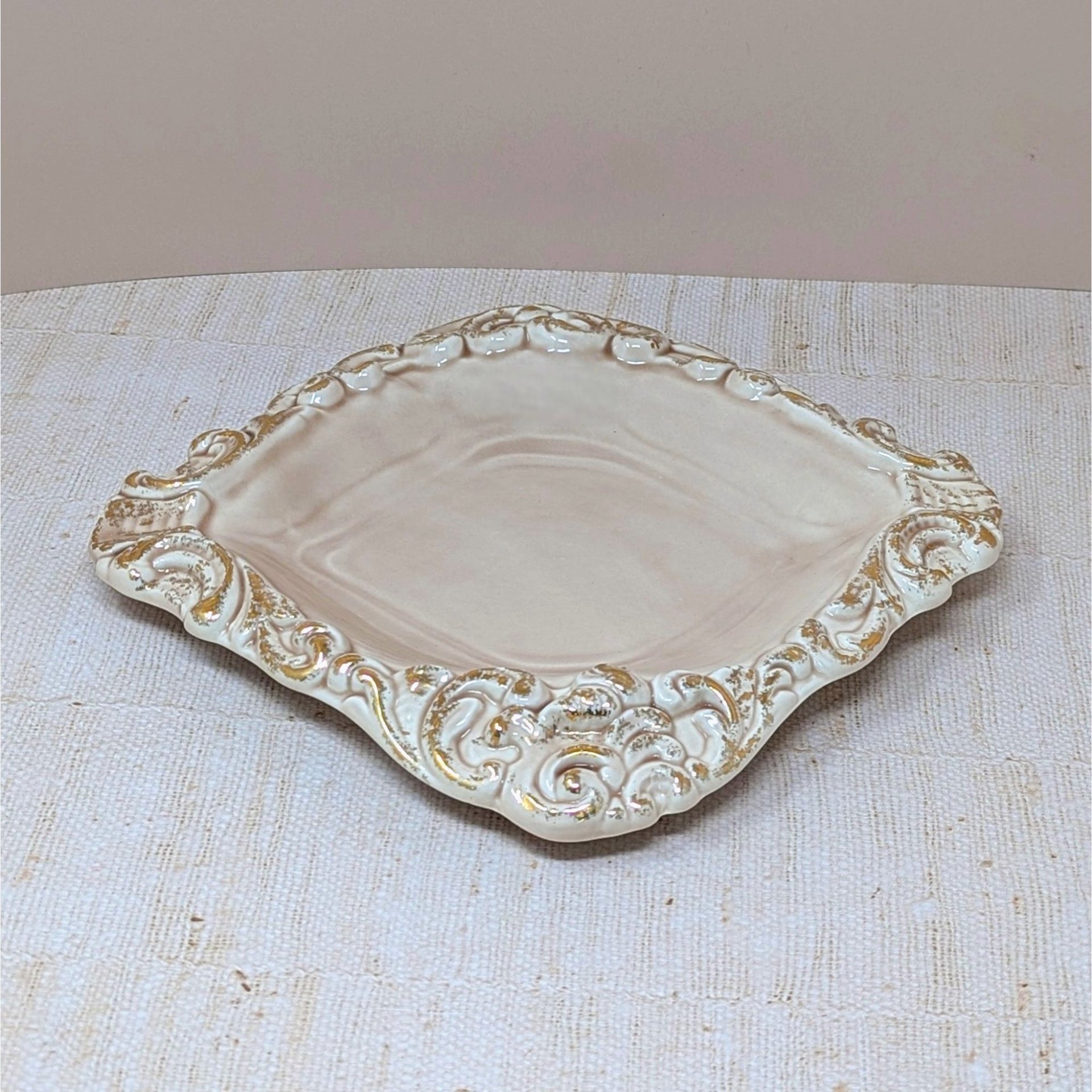 A square pink ceramic ashtray with a glossy finish. The edges are scalloped with embossed scroll patterns highlighted in distressed gold paint.
