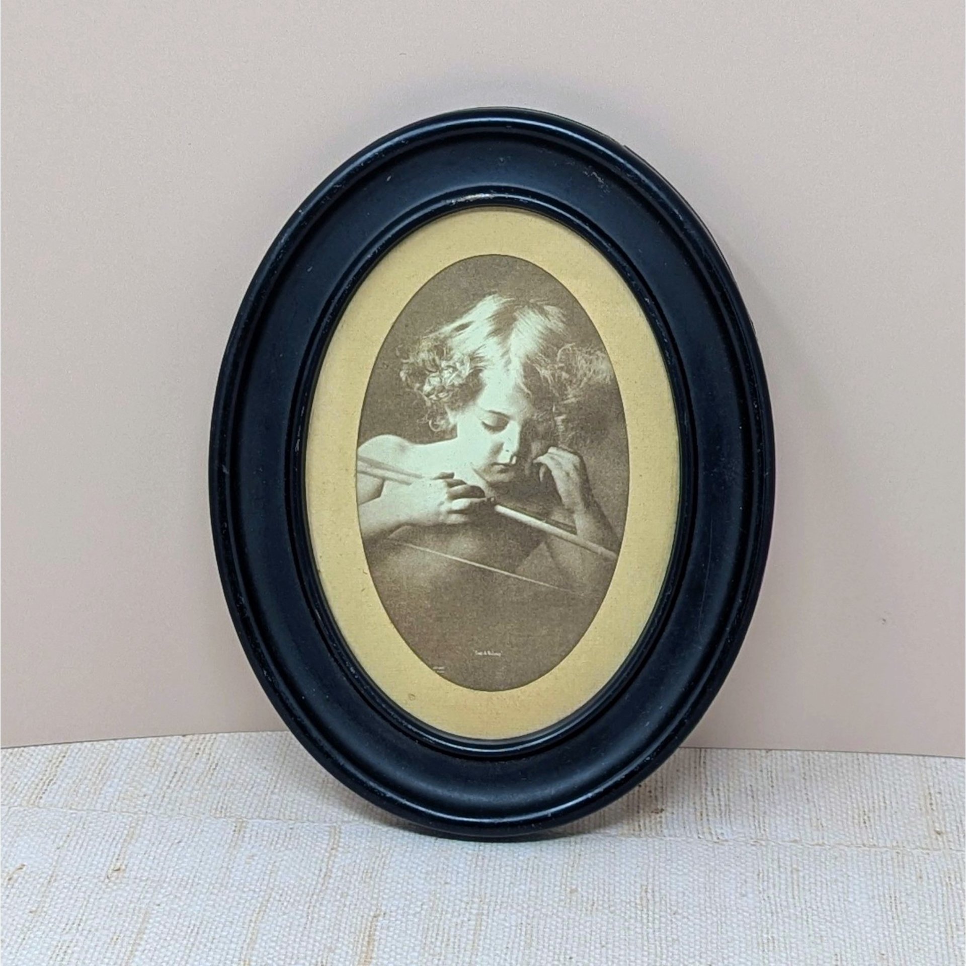 An oval vintage sepia-toned print titled "Cupid Asleep" by M.B. Parkinson. It depicts a young child with curls leaning over a bow. The print is housed in a thick, matte black wooden oval frame. 8.5 by 6.75 inches.A vintage sepia-toned oval print of M.B. Parkinson's "Cupid Asleep" featuring a young child with wings holding a bow. Housed in a black oval wooden frame with visible surface scrapes and aging.