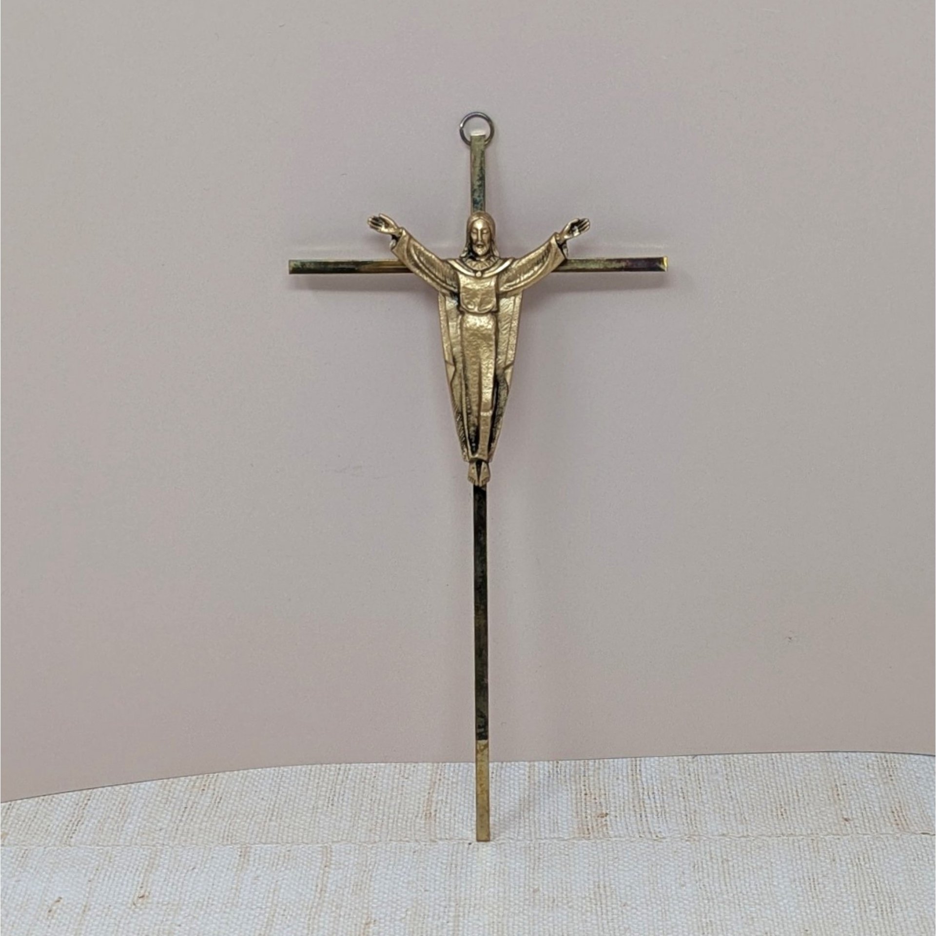 A vertical 10-inch brass crucifix wall hanging. Features a slender, modernist cross with a sculptural figure of the Risen Christ with arms outstretched. Aged brass finish with authentic patina.
