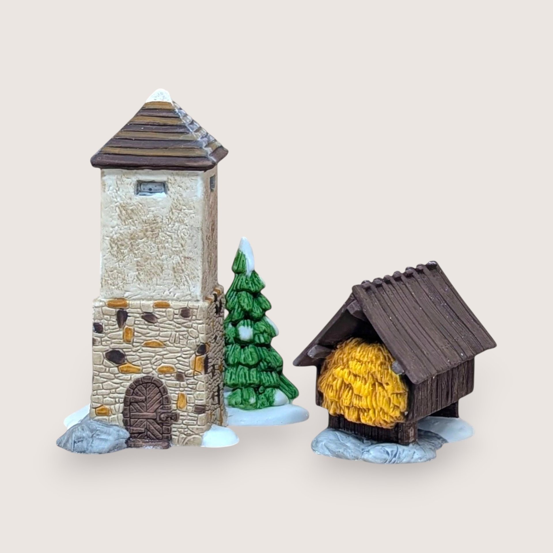 Miniature village stone tower, snow-capped evergreen tree, and a small wooden stable filled with yellow hay on a neutral background.