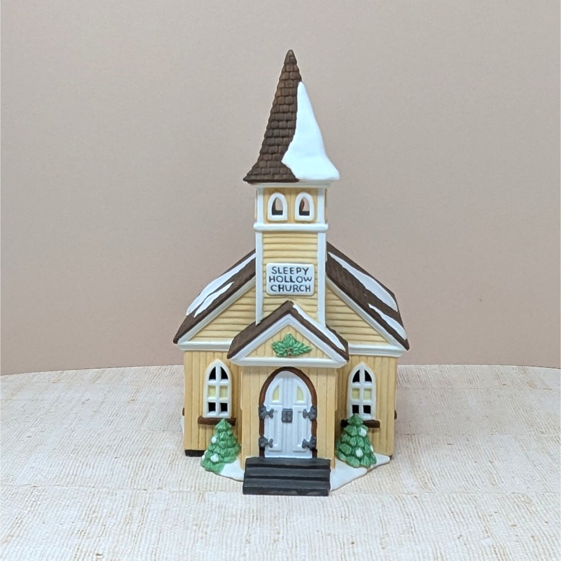 A vertical 9-inch hand-painted porcelain church with a tall steeple and creamy yellow siding. Features "Sleepy Hollow Church" signage, snow-dusted roofs, and small green evergreens at the entrance. Topped with a brown shingled spire.