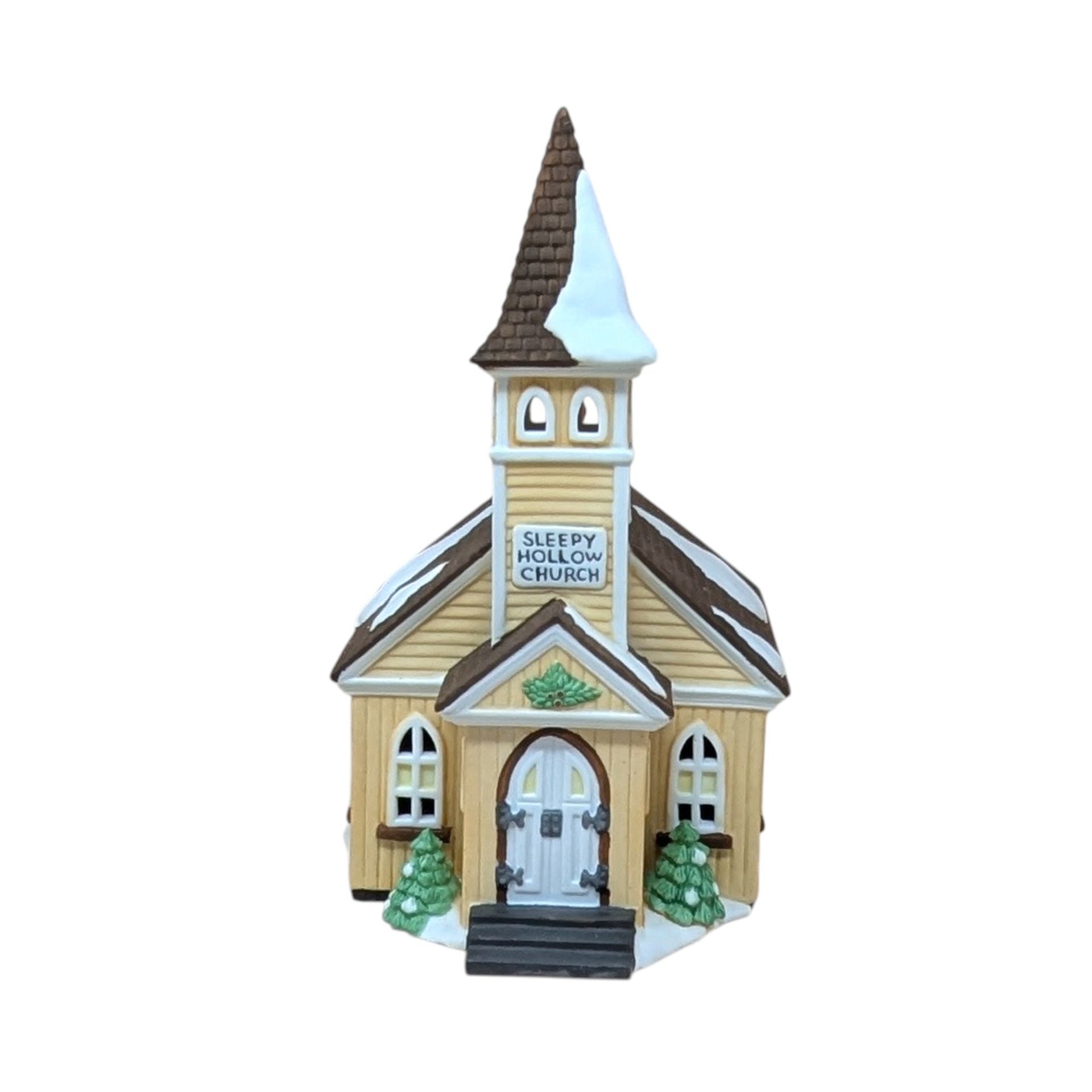 A vertical 9-inch hand-painted porcelain church with a tall steeple and creamy yellow siding. Features "Sleepy Hollow Church" signage, snow-dusted roofs, and small green evergreens at the entrance. Topped with a brown shingled spire.