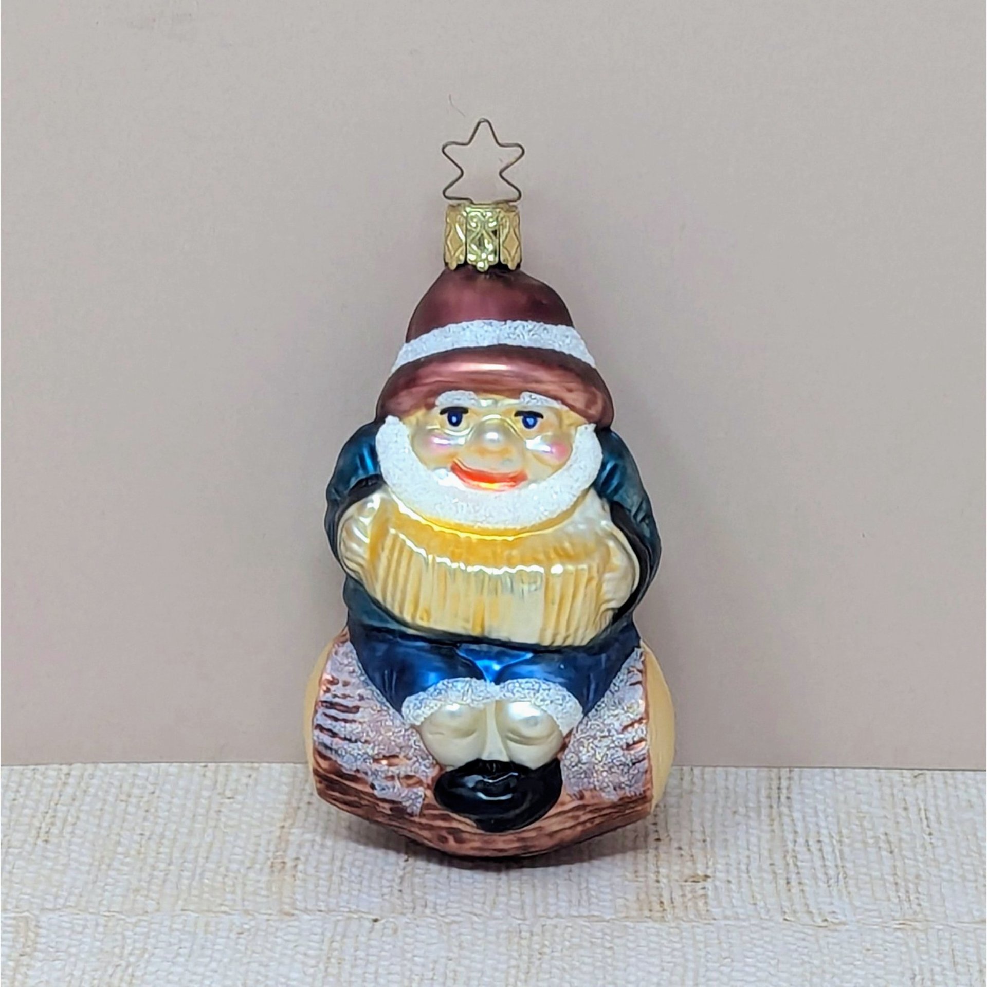 A hand-blown glass ornament of a cheerful gnome with a white beard and red hat, sitting on a brown log and playing a gold accordion. Features a gold star-shaped hanging cap. 4.25 inches tall