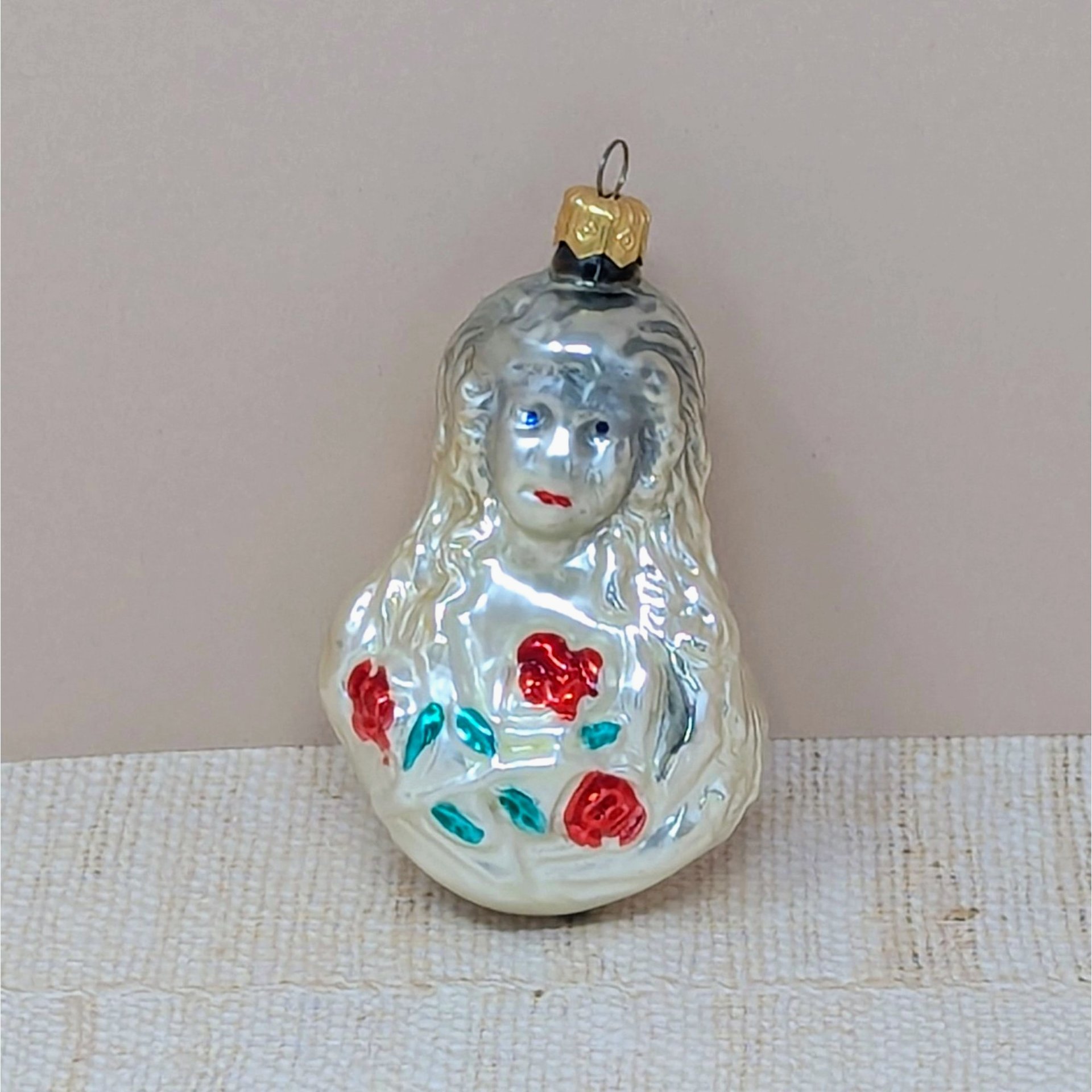 A vintage mercury glass ornament featuring a Victorian-style girl with long, flowing hair holding a bouquet of red roses with green leaves. The glass has a warm, milky-white luster with hand-painted facial details.