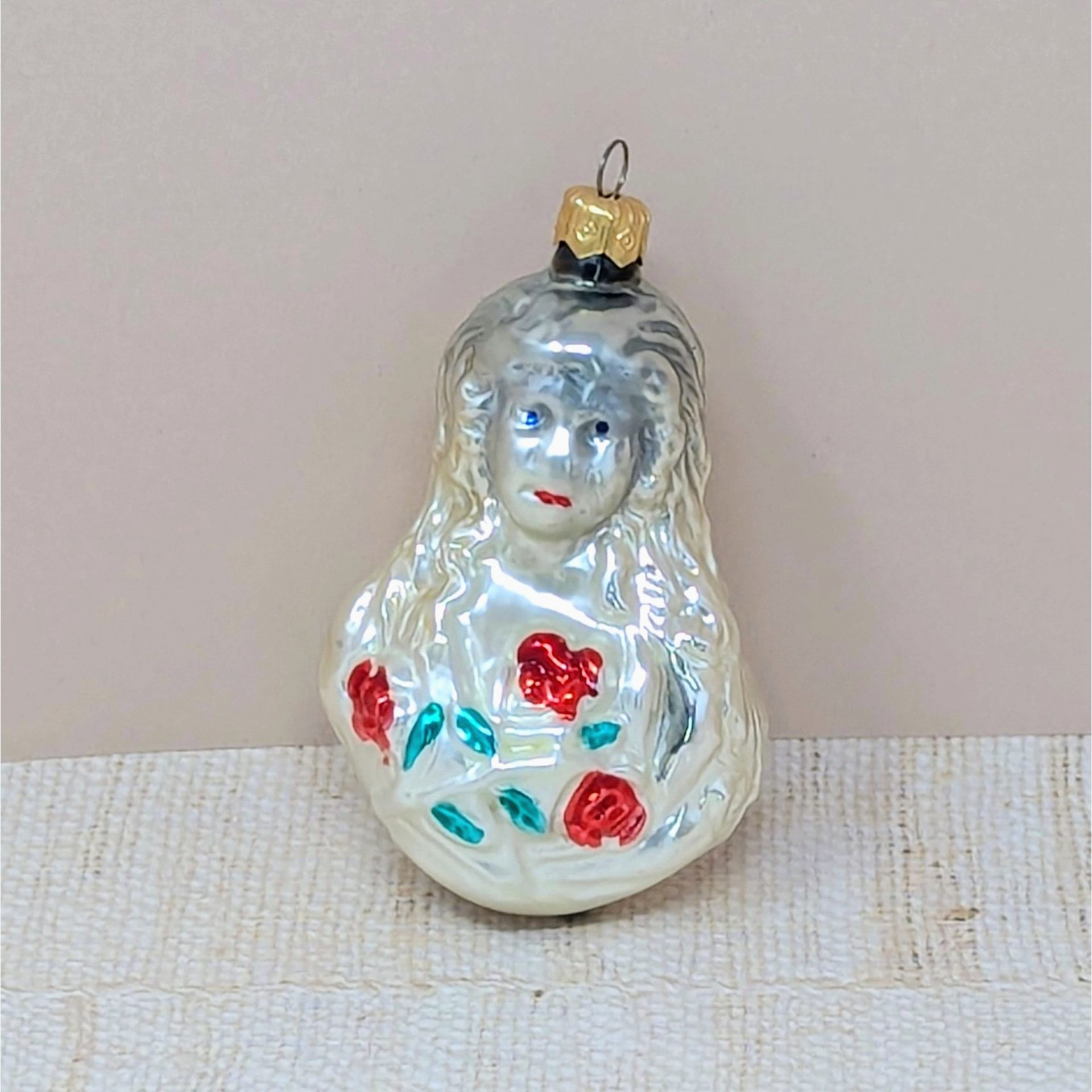 A vintage mercury glass ornament featuring a Victorian-style girl with long, flowing hair holding a bouquet of red roses with green leaves. The glass has a warm, milky-white luster with hand-painted facial details.