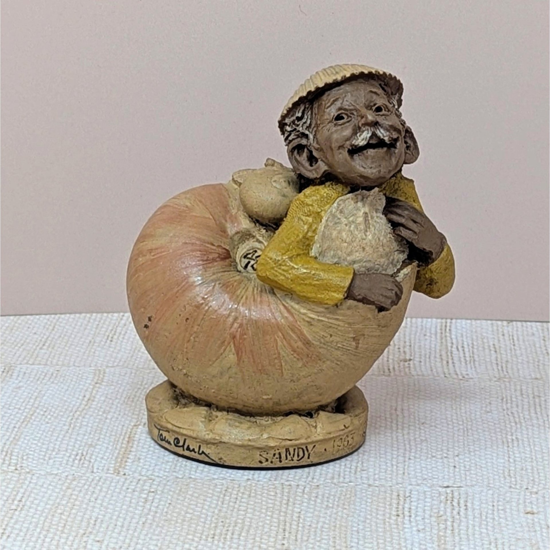 A hand-cast resin sculpture of a smiling gnome-like character named Sandy emerging from a large peach-colored seashell. The character wears a yellow jacket and a small hat. 4.75 inches tall.