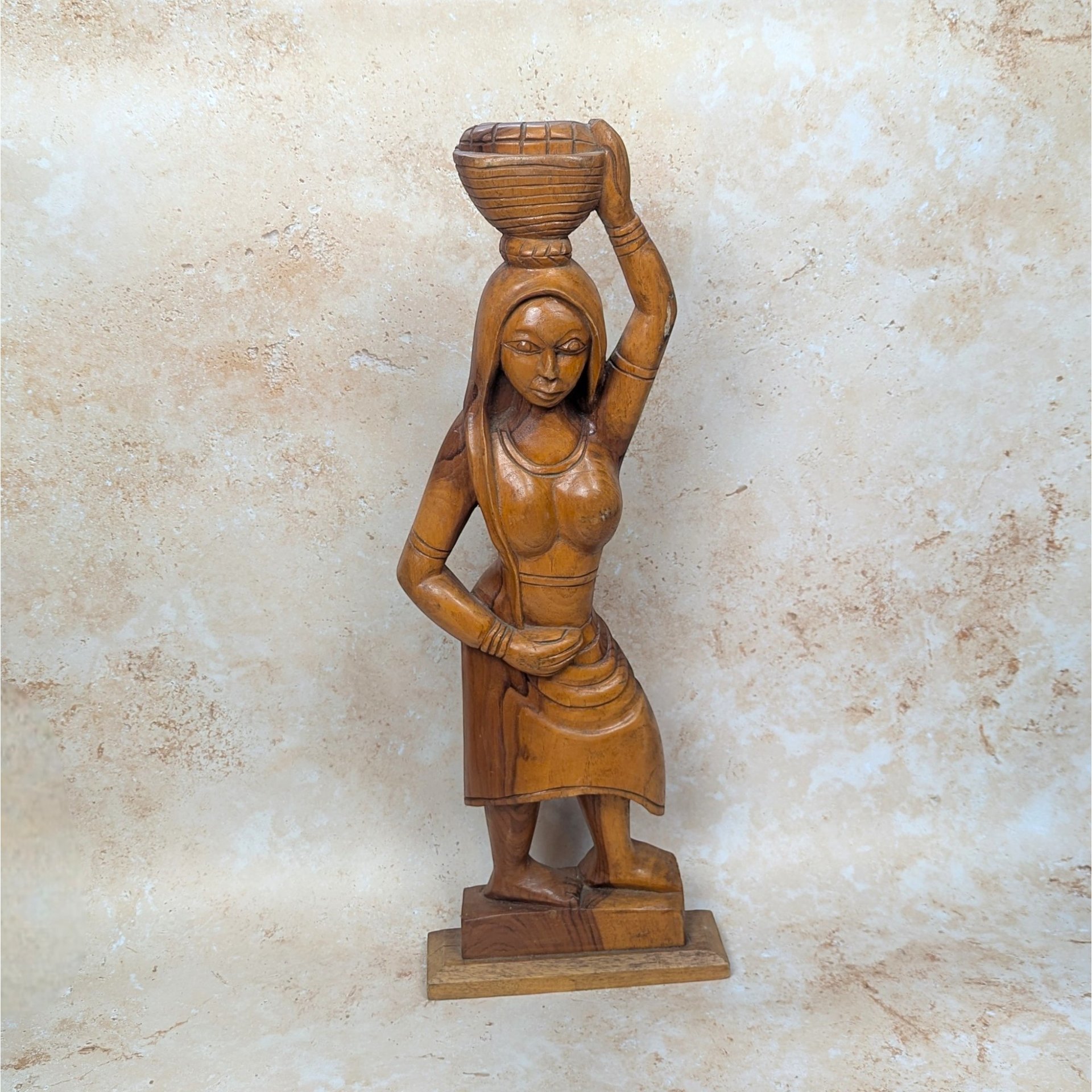 A vintage mid-century hand carved wooden statue of an African woman on a tan textured background. Standing twenty four inches tall, the solid timber figure gracefully balances a woven basket on her head with one arm raised.