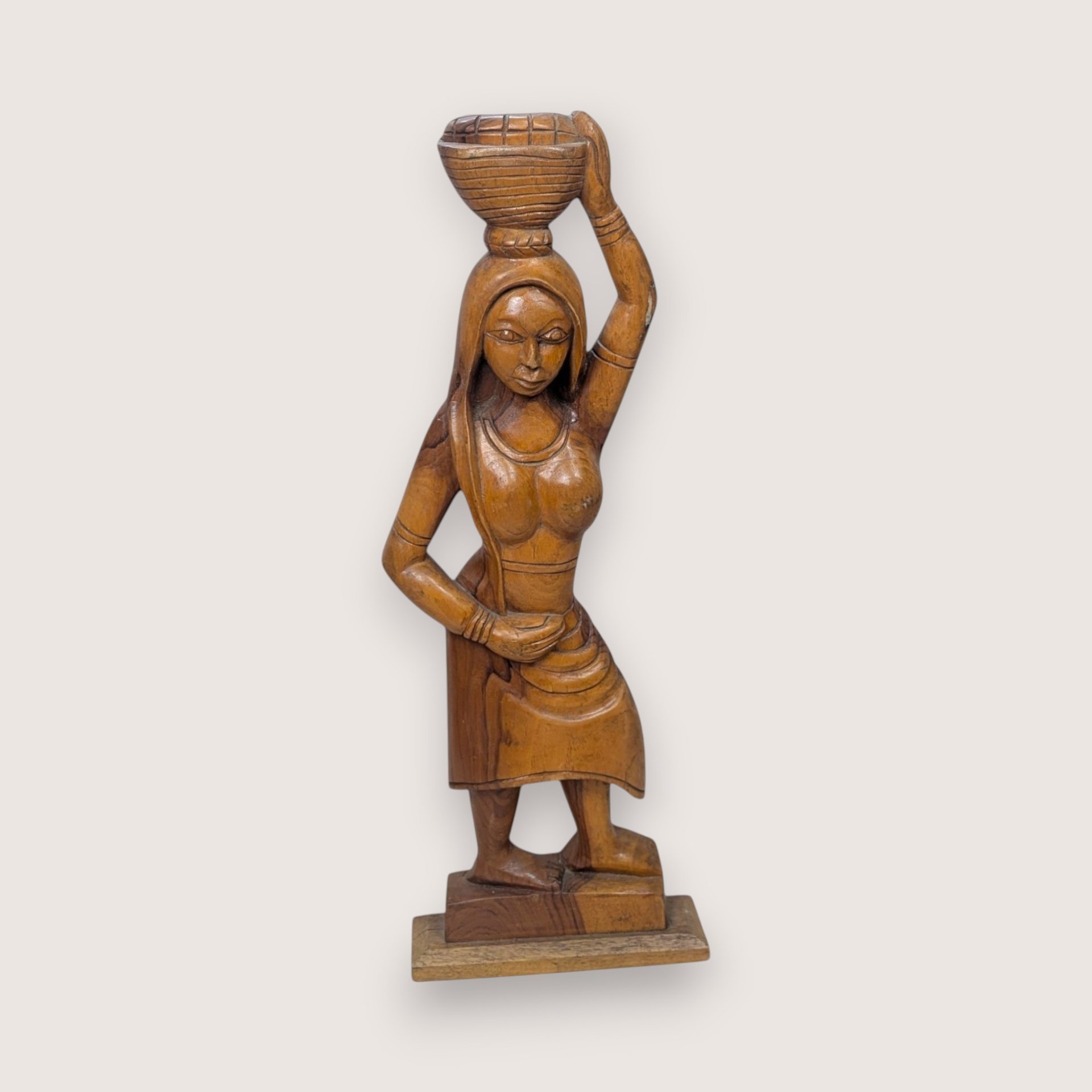 Carved wooden statue of a woman carrying a woven basket on her head, isolated on a light neutral background.