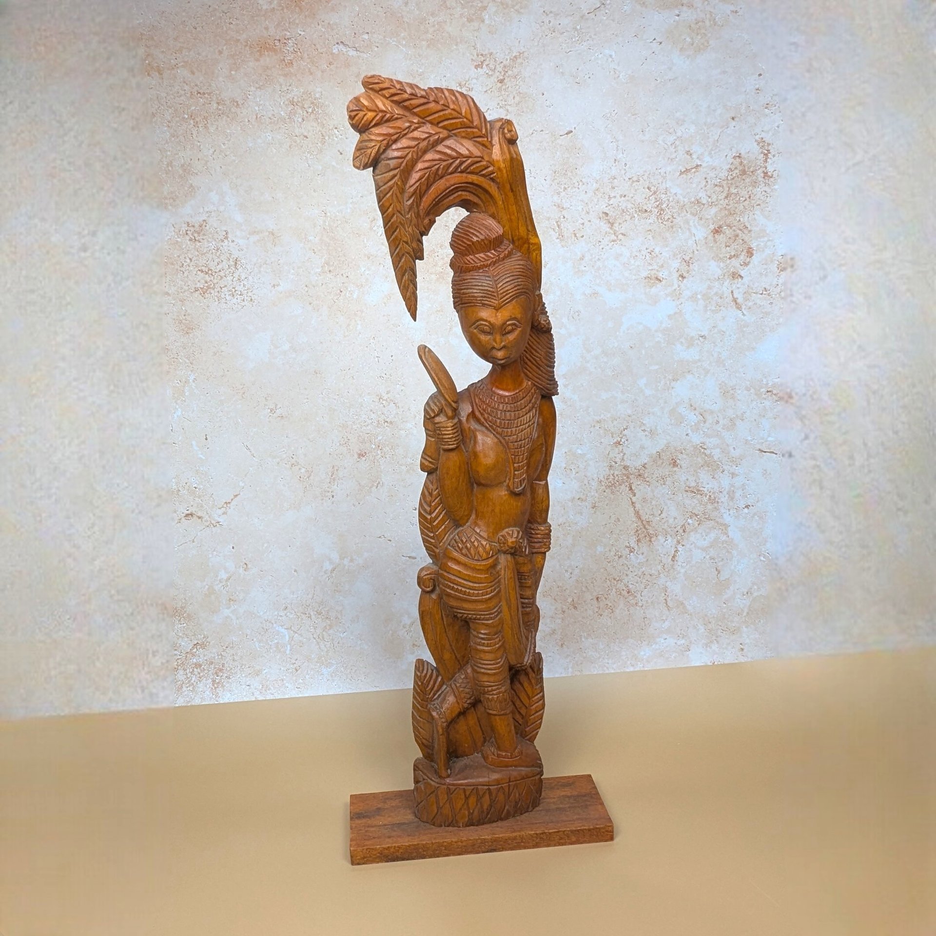 A vintage mid-century hand carved wooden statue of a Balinese woman on a tan textured background. Standing twenty eight inches tall, the highly detailed timber figure poses gracefully beneath an ornate, stylized palm frond canopy.