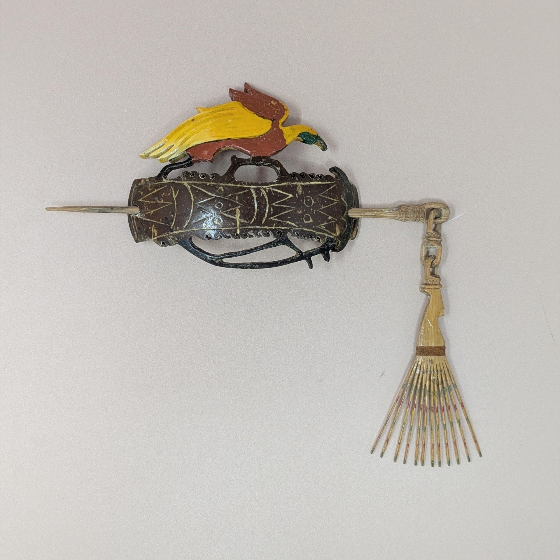 A hand-carved wooden hair slide featuring a red and yellow bird perched atop an ornate brown base with geometric carvings. A wooden pin with a dangling tassel secures it. 4 inches wide.