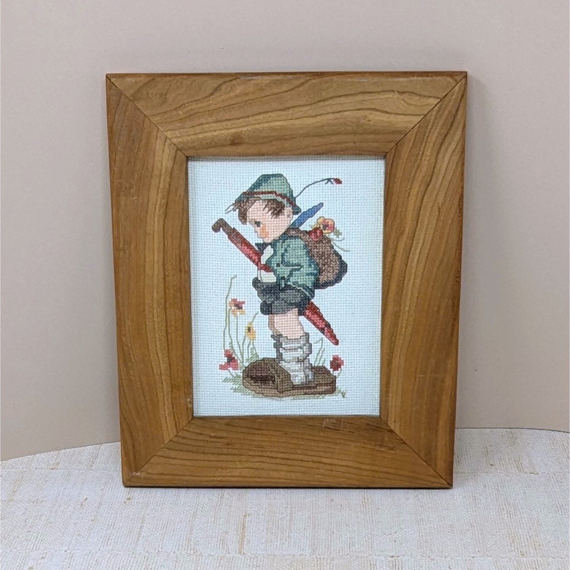 A completed and framed cross-stitch of a young boy in traditional Alpine clothes holding a large red umbrella, known as the Hummel "It Looks Like Rain" design. 11 by 9 inches.