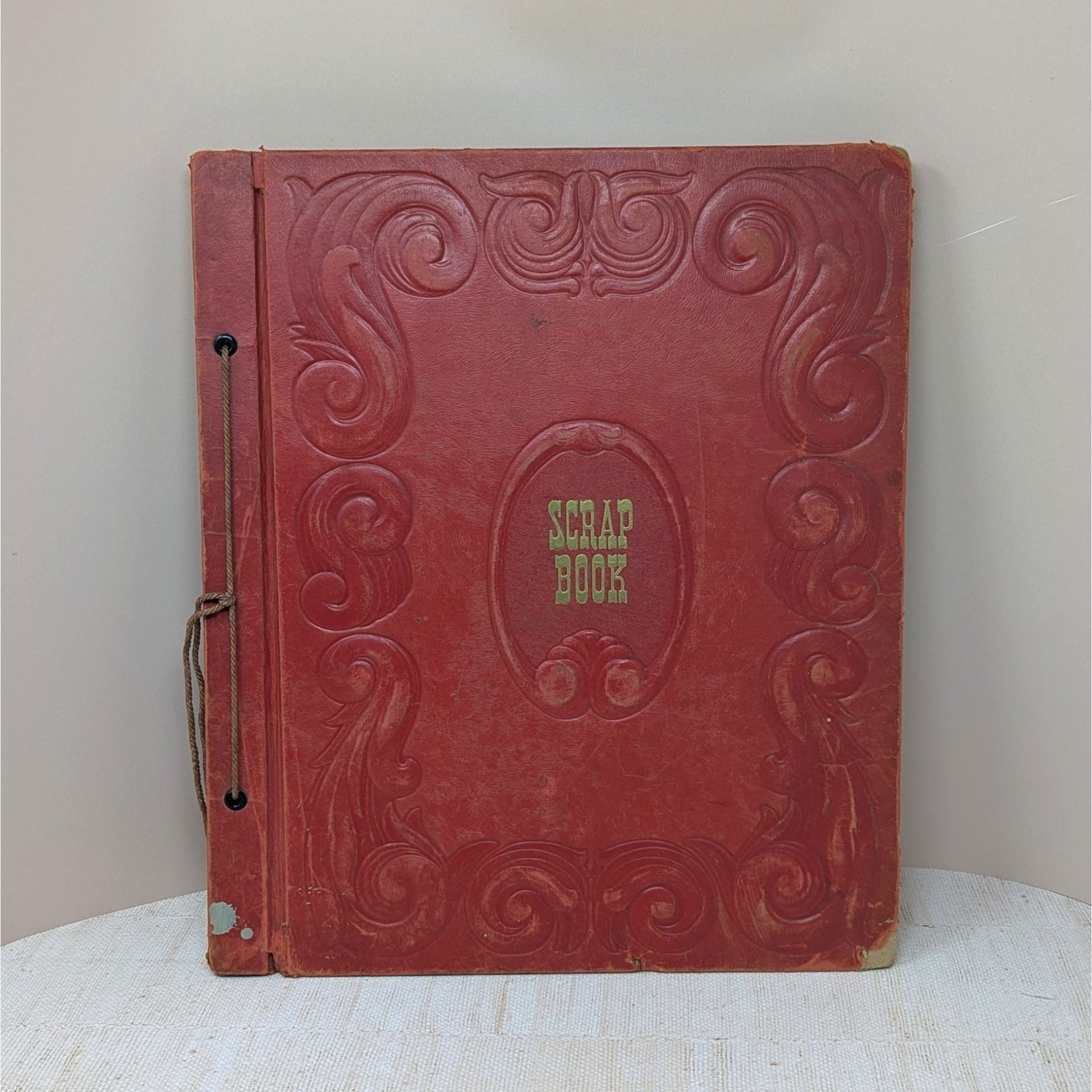 A large, vintage red leatherette scrapbook with an ornate embossed border and "Scrapbook" in gold lettering at the center. The cover shows significant age-related wear and is secured by a brown cord binding.