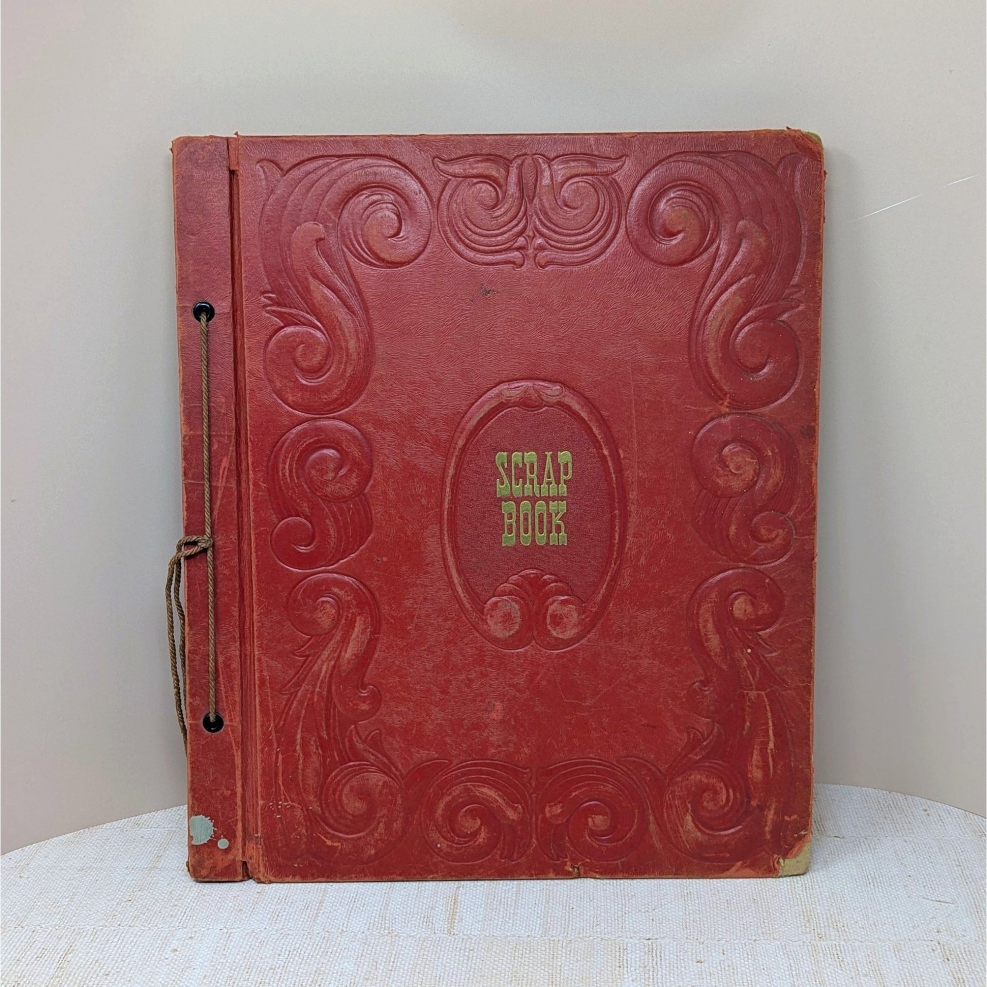 A large, vintage red leatherette scrapbook with an ornate embossed border and "Scrapbook" in gold lettering at the center. The cover shows significant age-related wear and is secured by a brown cord binding.