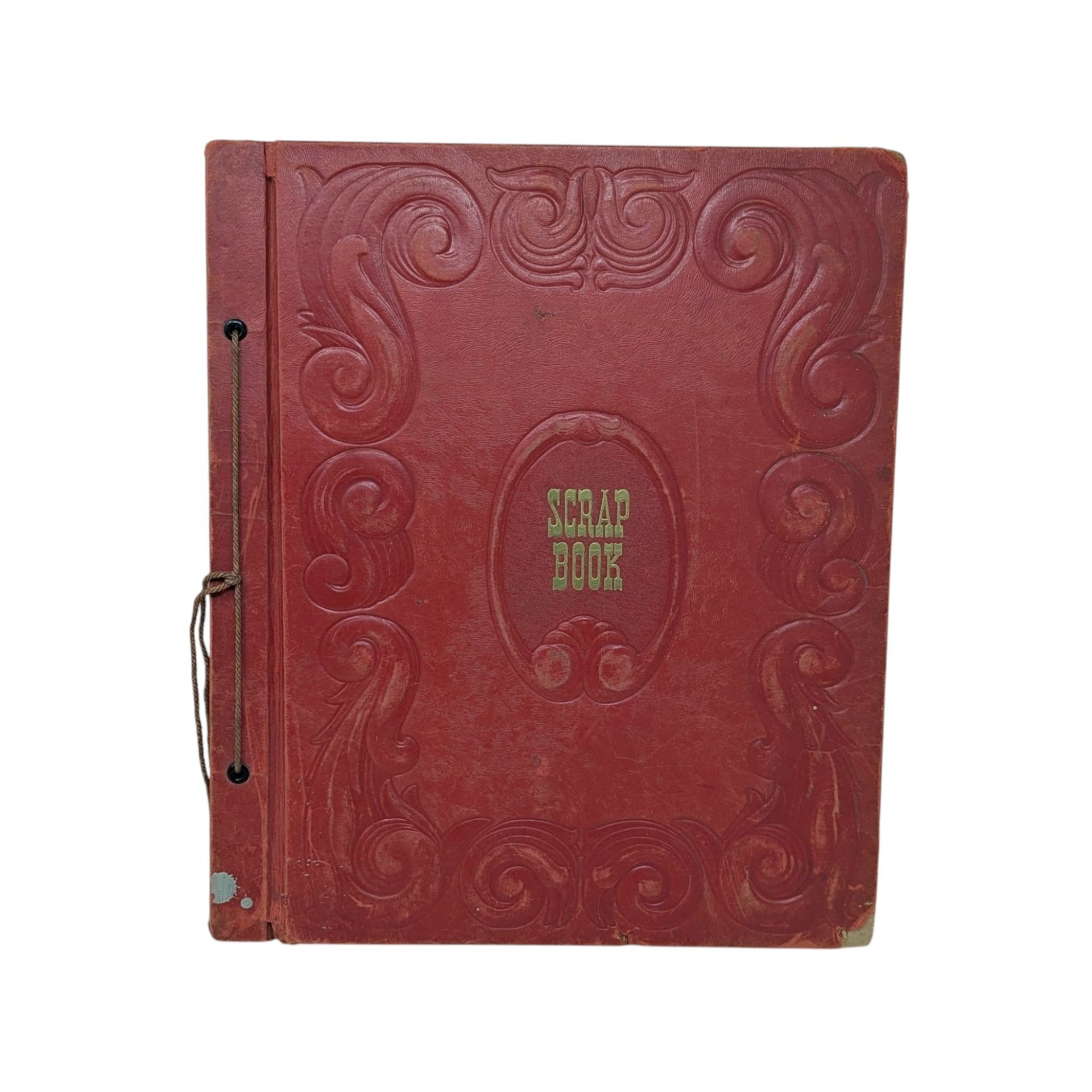 A large, vintage red leatherette scrapbook with an ornate embossed border and "Scrapbook" in gold lettering at the center. The cover shows significant age-related wear and is secured by a brown cord binding.