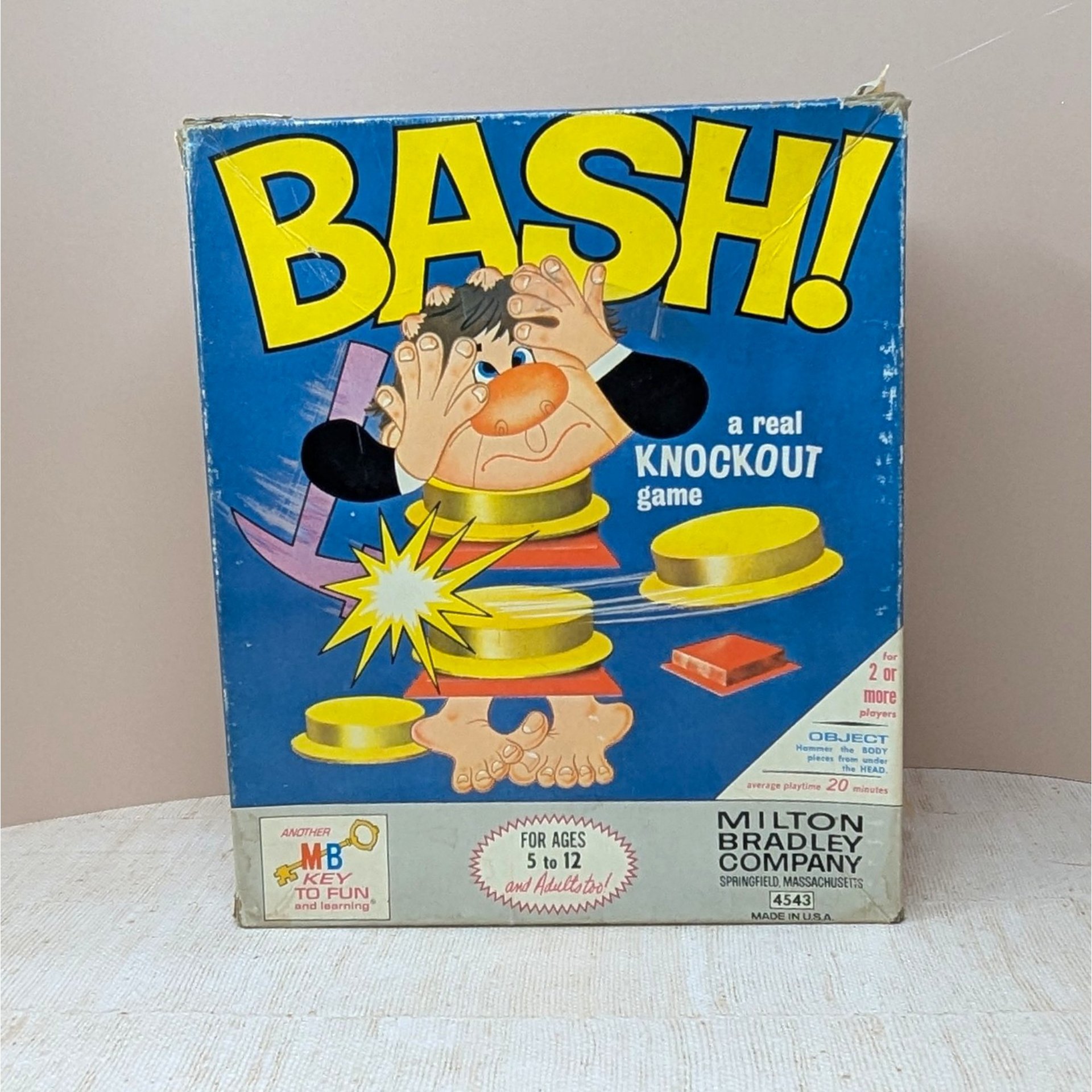 The original 1965 box for the Milton Bradley game "BASH!" featuring a bright blue background and a cartoon man being "bashed" apart. The box shows vintage shelf wear but remains structurally sound with vibrant yellow lettering.