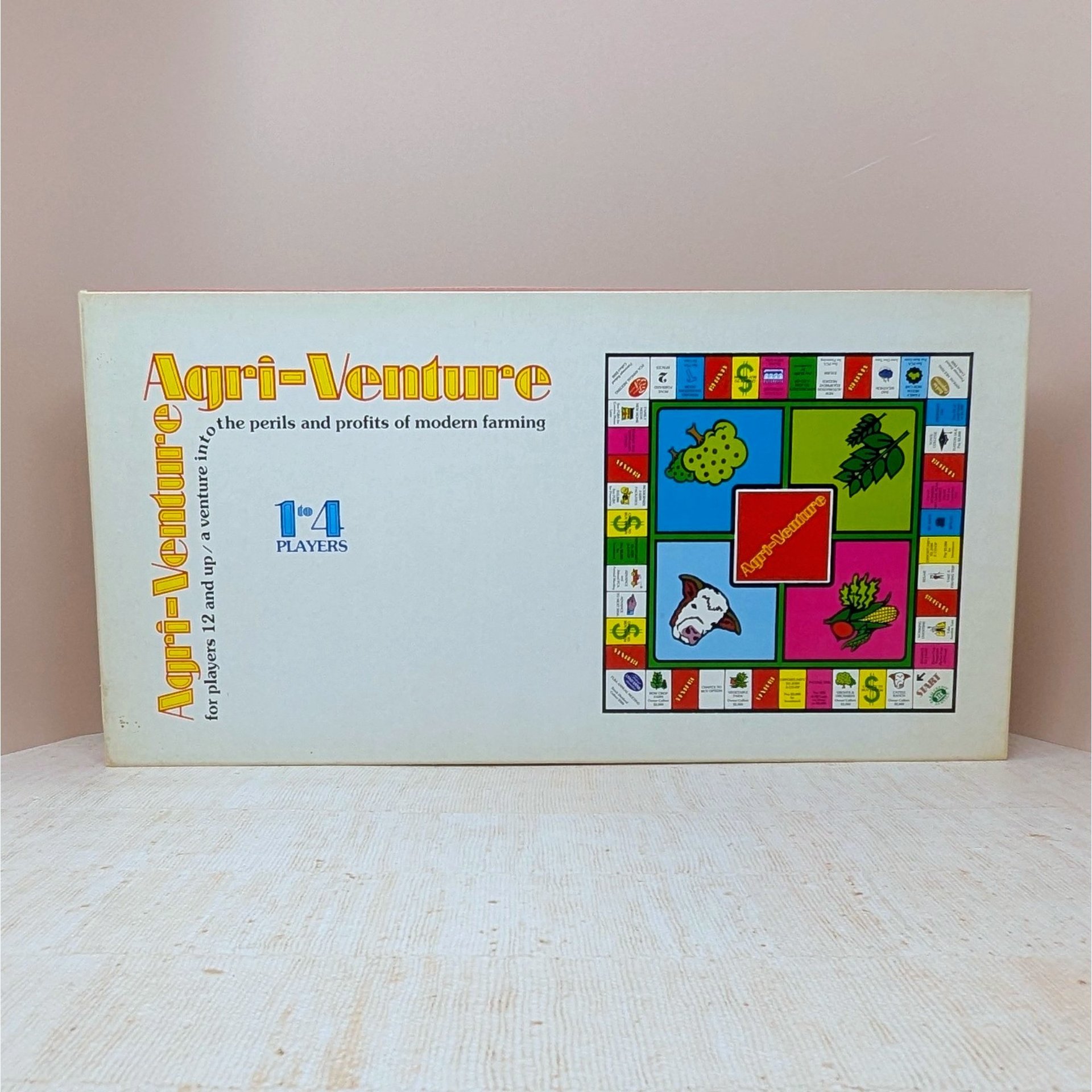 A long white rectangular game box for Agri-Venture. The text is in a retro orange and yellow serif font. A colorful graphic of the square game board is on the right, featuring illustrations of a cow, corn, and trees.