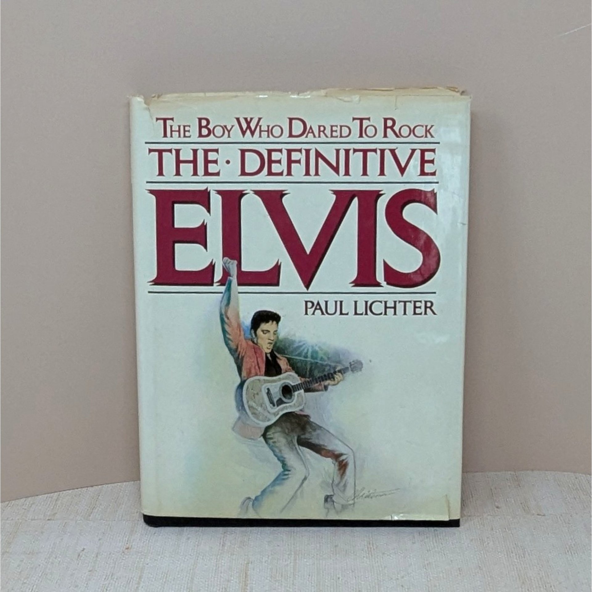A large hardcover book titled The Boy Who Dared To Rock: The Definitive Elvis. The white dust jacket features an illustration of Elvis Presley in a pink jacket with a guitar. The jacket shows visible edge wear and small tears.