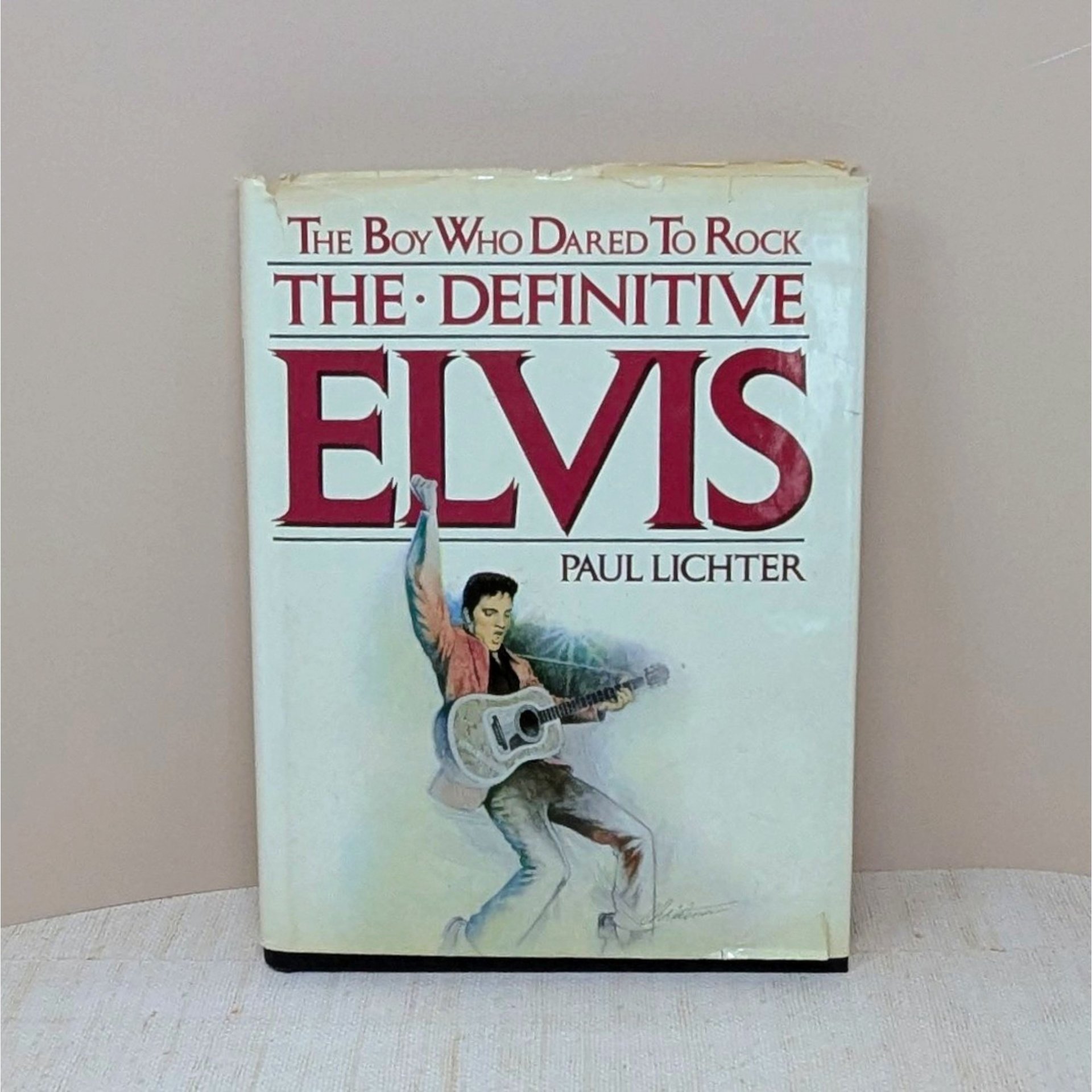 A large hardcover book titled The Boy Who Dared To Rock: The Definitive Elvis. The white dust jacket features an illustration of Elvis Presley in a pink jacket with a guitar. The jacket shows visible edge wear and small tears.