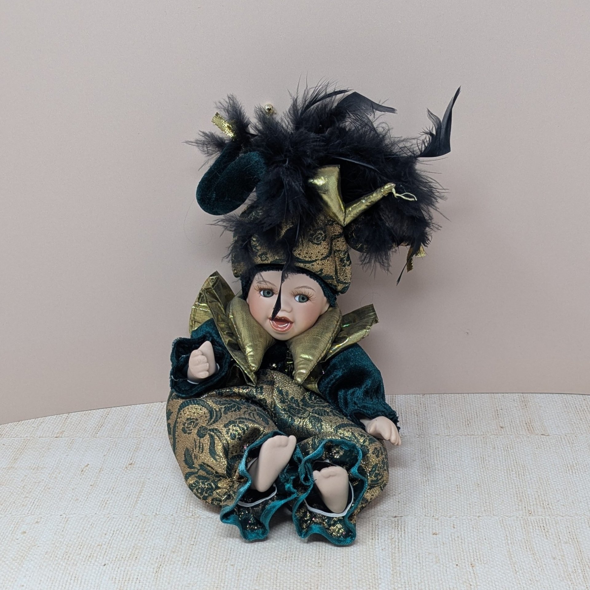 A 12-inch porcelain baby doll dressed as a jester in emerald green velvet and gold brocade, featuring a large feathered jester hat and gold metallic ruff.
