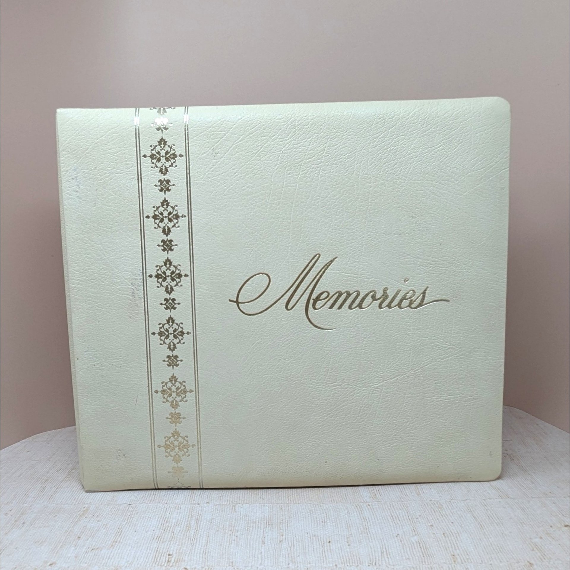 A large, square vintage photo album in cream faux leather. The word Memories is printed in a gold cursive script on the front. A decorative vertical border of gold filigree crosses runs down the left side.