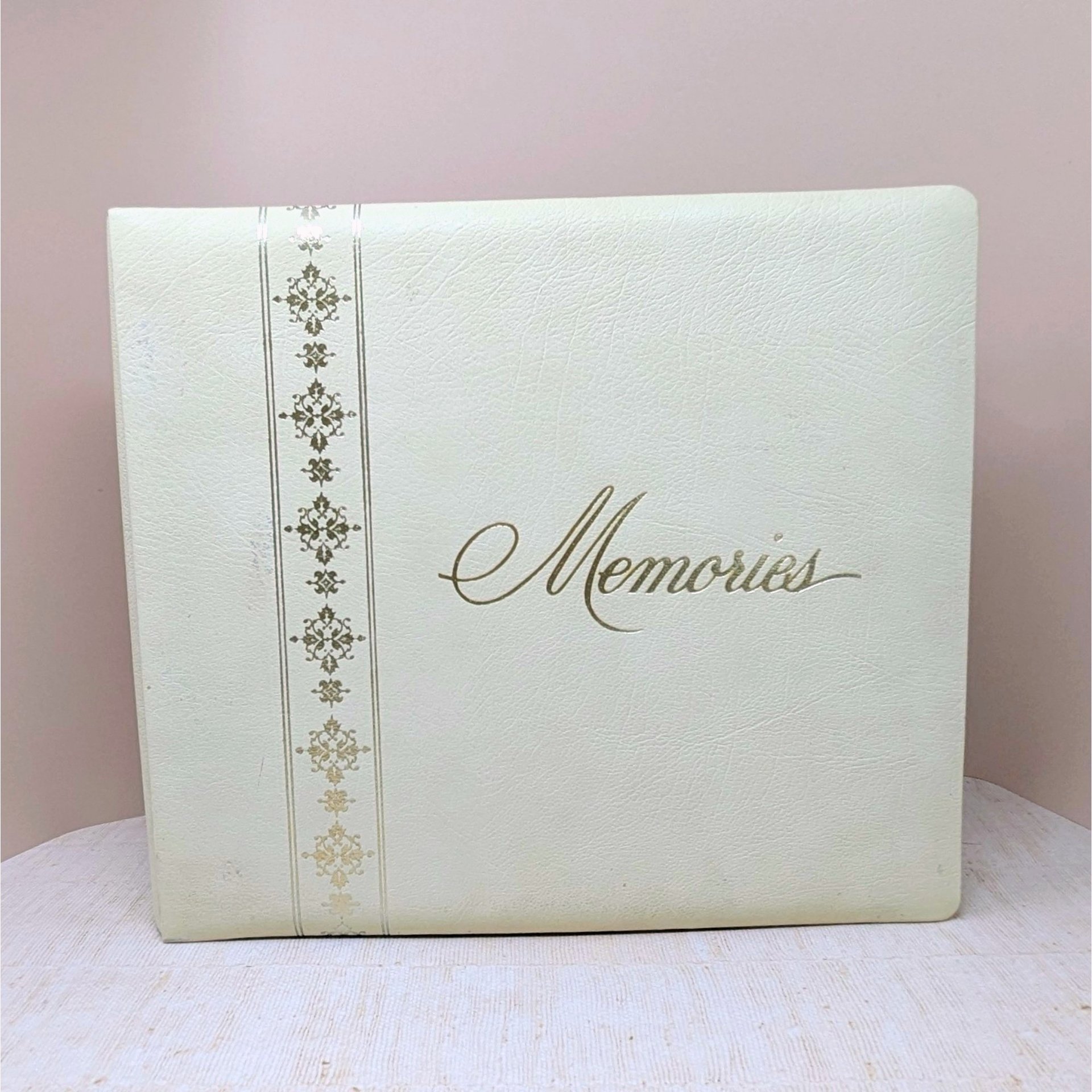 A large, square vintage photo album in cream faux leather. The word Memories is printed in a gold cursive script on the front. A decorative vertical border of gold filigree crosses runs down the left side.