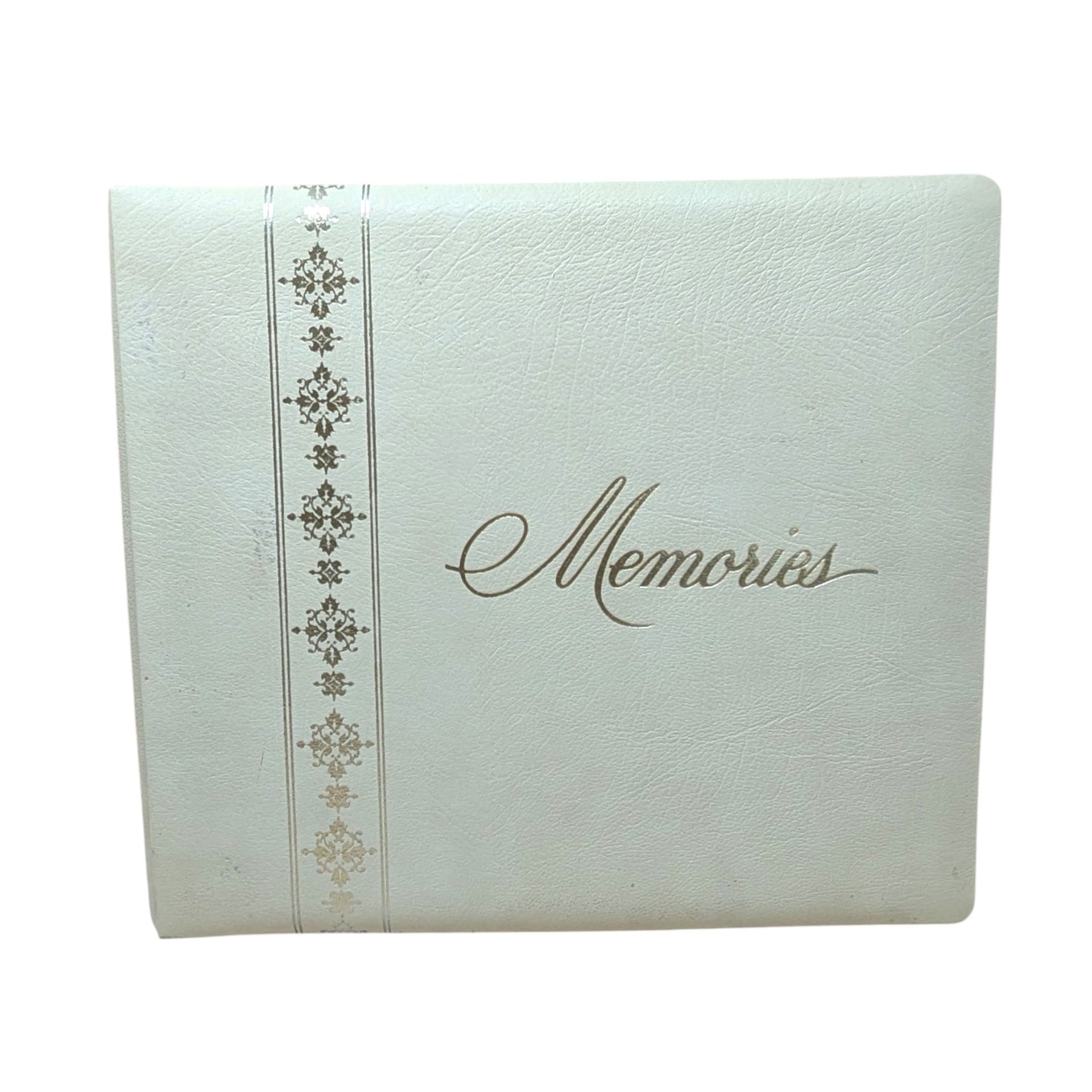 A large, square vintage photo album in cream faux leather. The word Memories is printed in a gold cursive script on the front. A decorative vertical border of gold filigree crosses runs down the left side.