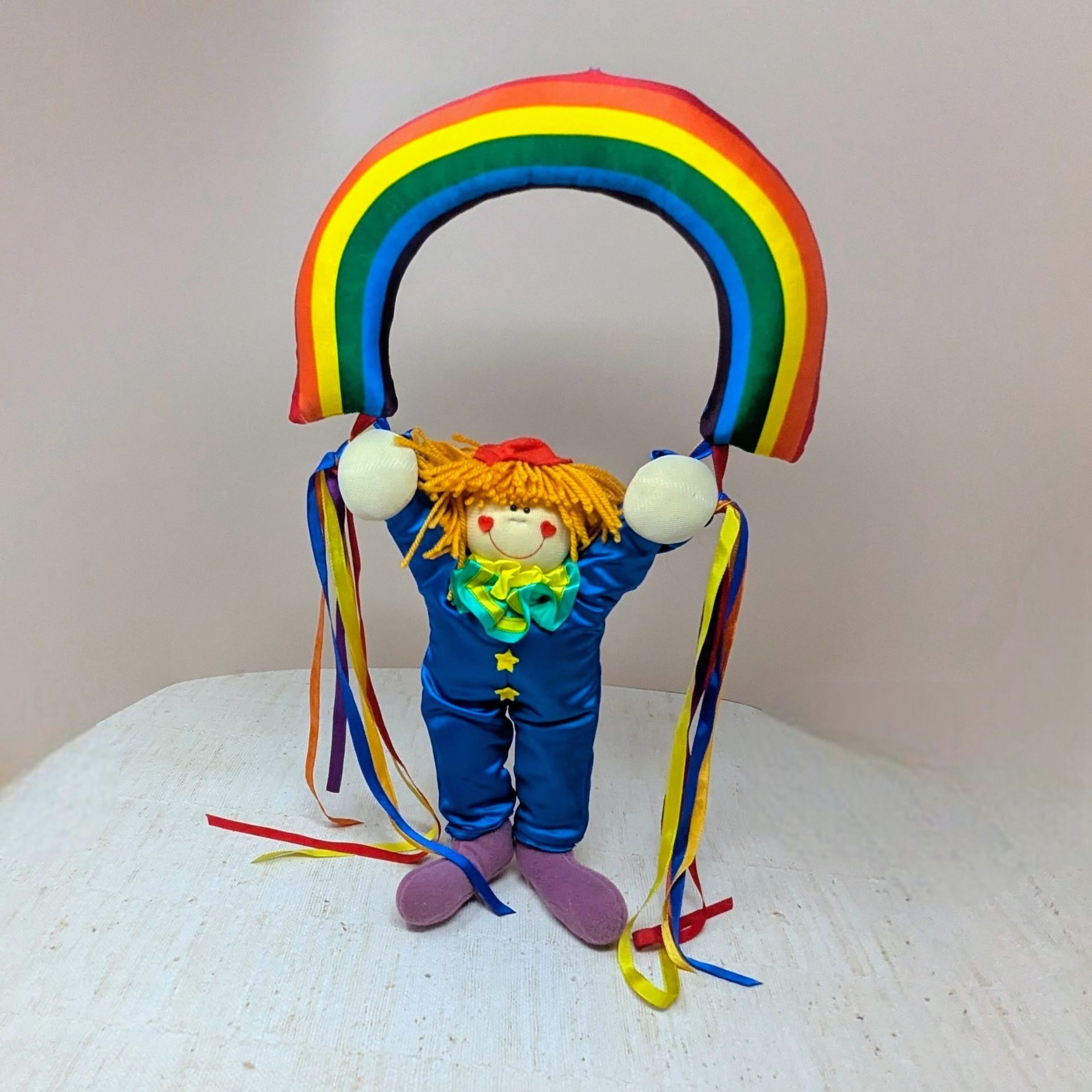 A 1984 vintage Dakin plush wall hanging featuring a small clown with yellow yarn hair and a blue satin suit. The clown is holding up a large stuffed rainbow arch. Long colorful ribbons hang from the clown's hands.