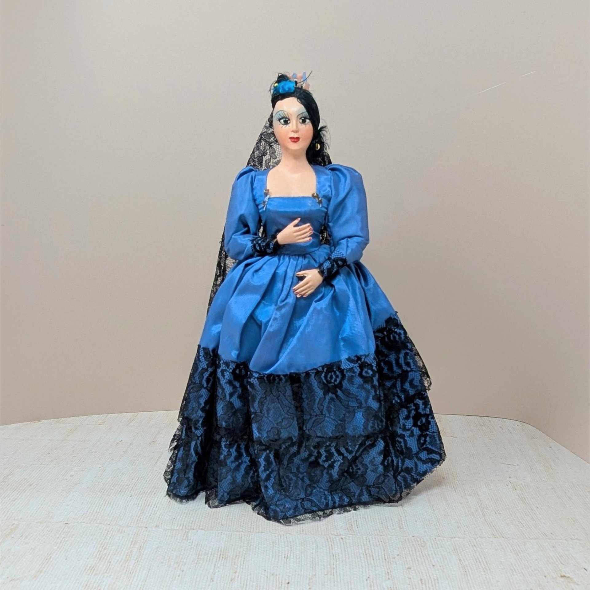 A vintage 13-inch Spanish souvenir doll featuring a cobalt blue satin dress with black lace tiers, a matching black lace mantilla veil, and dramatic hand-painted facial features against a neutral background.