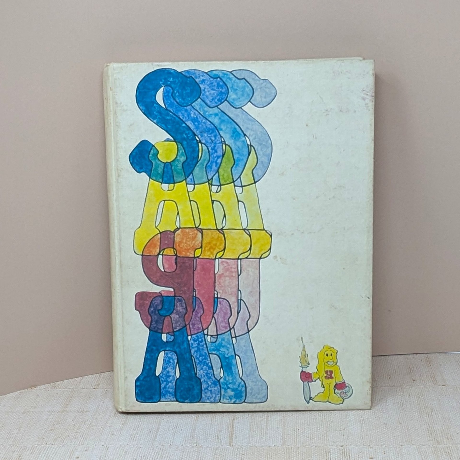 A close-up of a 1975 high school yearbook cover with a bold, multi-colored graphic design. The title SASH is layered in translucent, vibrant 70s typography over a dark background. The cloth texture of the hardcover is visible.