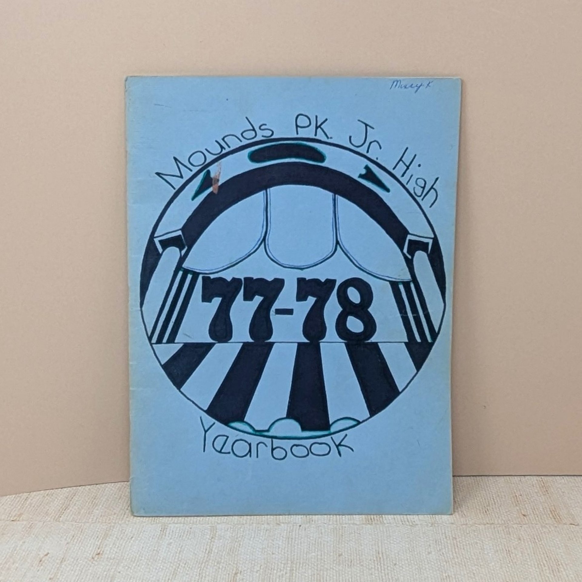 A vintage softcover yearbook featuring a light blue cover with a hand-drawn circular graphic in black ink. The center shows the bold numbers 77-78 under a theatrical or arched motif. A name is written in small ink at the top right.