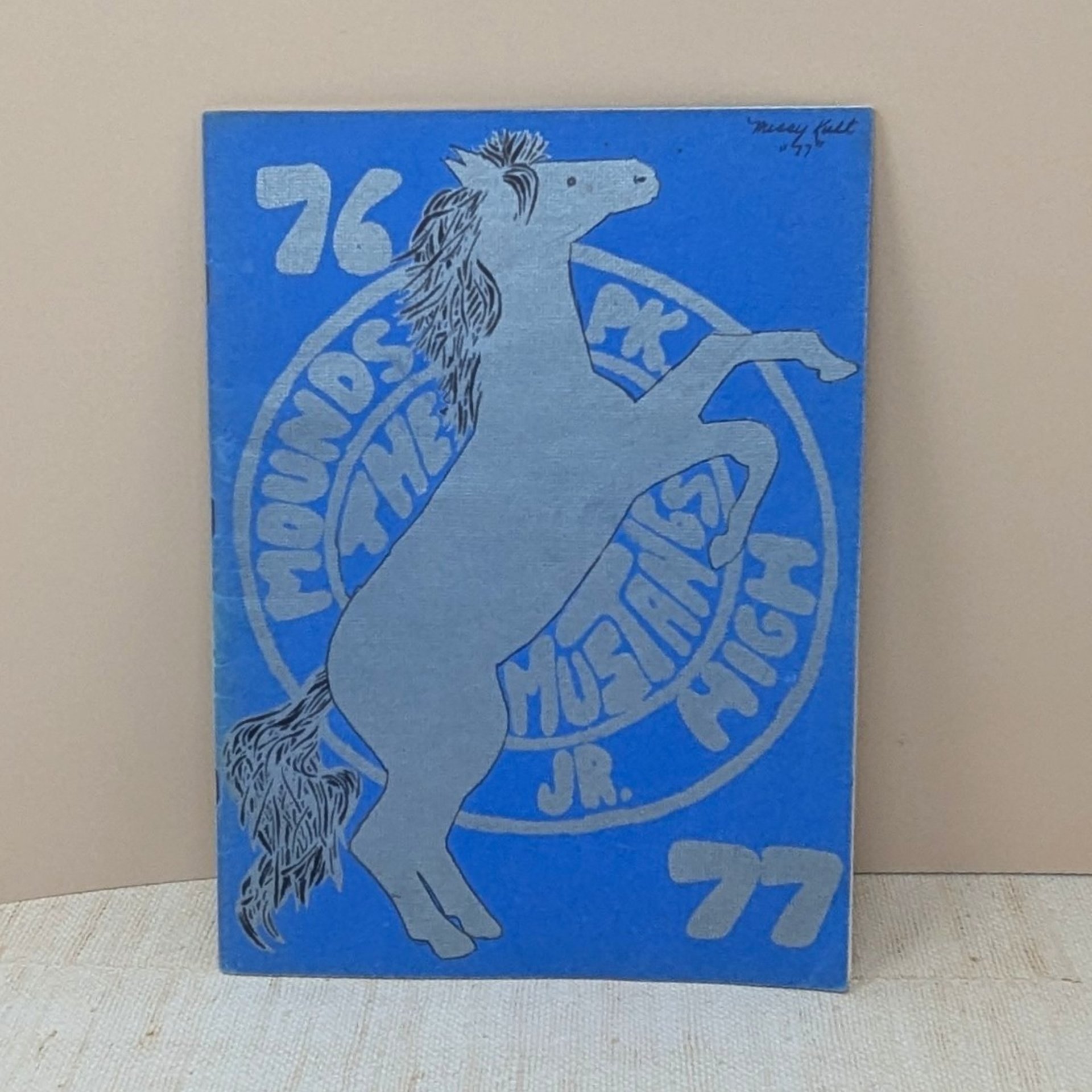 A vintage softcover yearbook with a bold blue cover. It features a large, silver-ink illustration of a rearing mustang horse centered over a circular school seal. The year 76-77 is printed in large block numbers.