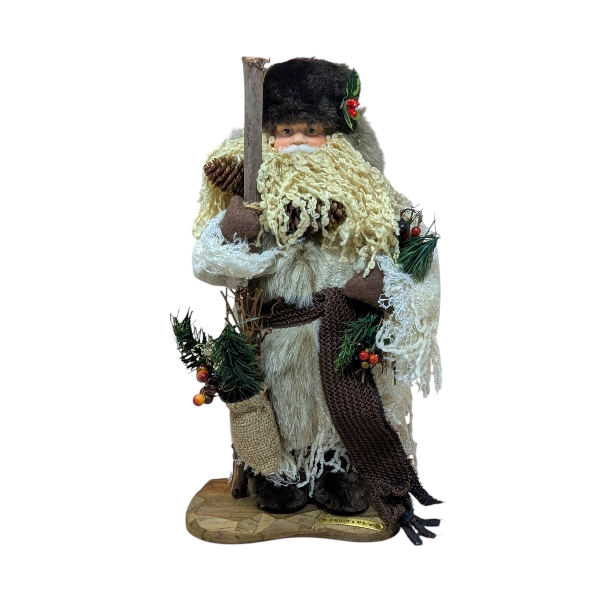 A tall, standing Father Christmas figure in a tan coat and brown fur hat. He has a long curly beard, holds a wooden staff, and carries a burlap sack with pine needles.