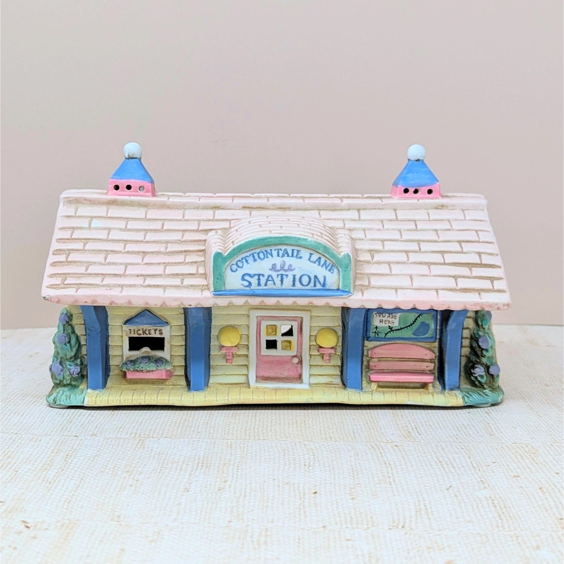 A vintage ceramic lighted house titled Cottontail Lane Station. It features a pink shingled roof with blue cupolas and a yellow clapboard exterior. The front includes a ticket window, a pink door, and a tiny bench under a "You Are Here" map.
