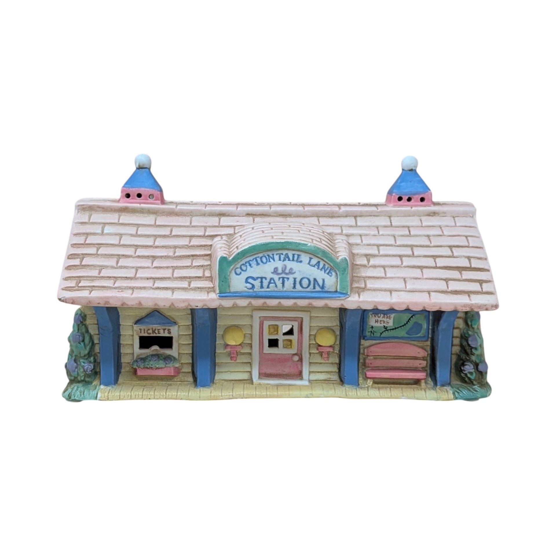 A vintage ceramic lighted house titled Cottontail Lane Station. It features a pink shingled roof with blue cupolas and a yellow clapboard exterior. The front includes a ticket window, a pink door, and a tiny bench under a "You Are Here" map.