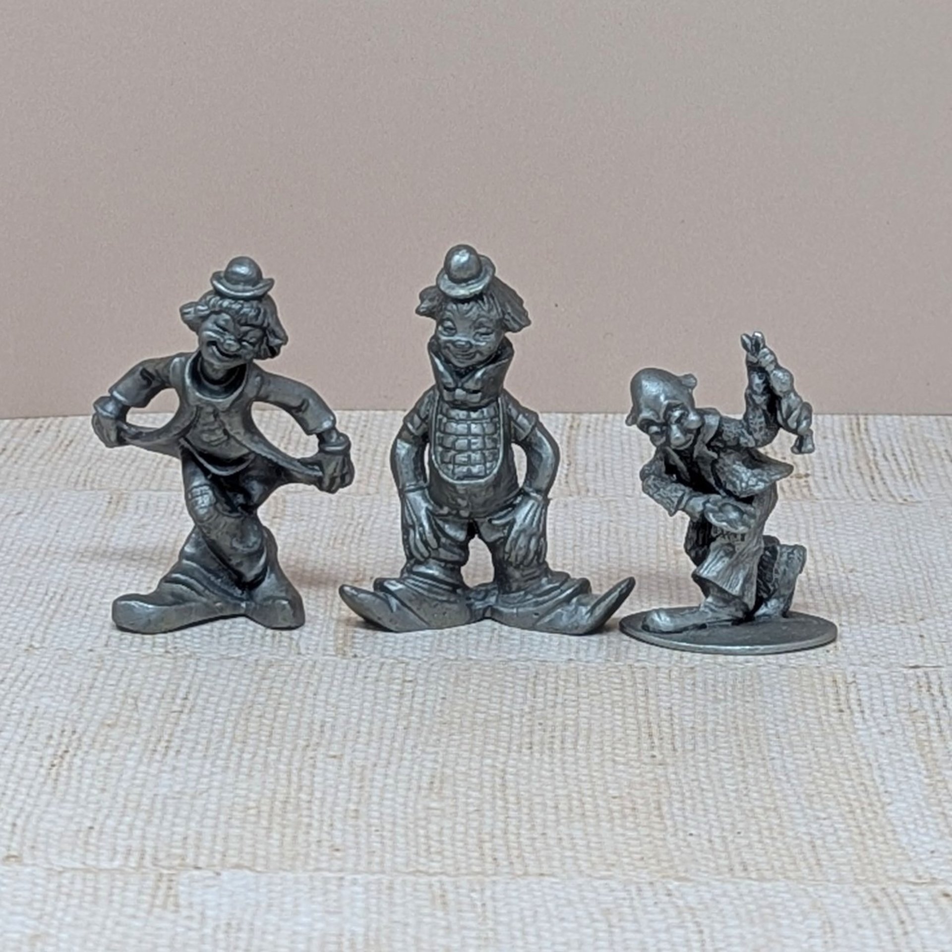 A trio of vintage silver-toned pewter clown figurines. One is in a deep curtsy, another is mid-stride in a classic circus pose, and the third is a dapper clown with a tall hat and buttons. Each figure has a matte metallic finish with fine sculptural details.