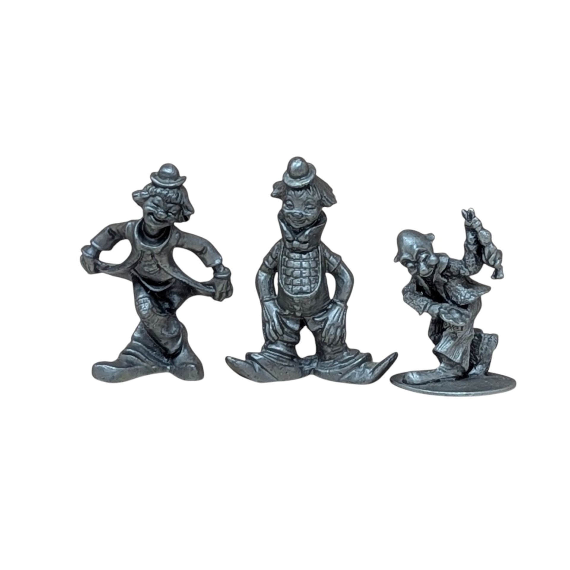 A trio of vintage silver-toned pewter clown figurines. One is in a deep curtsy, another is mid-stride in a classic circus pose, and the third is a dapper clown with a tall hat and buttons. Each figure has a matte metallic finish with fine sculptural details.