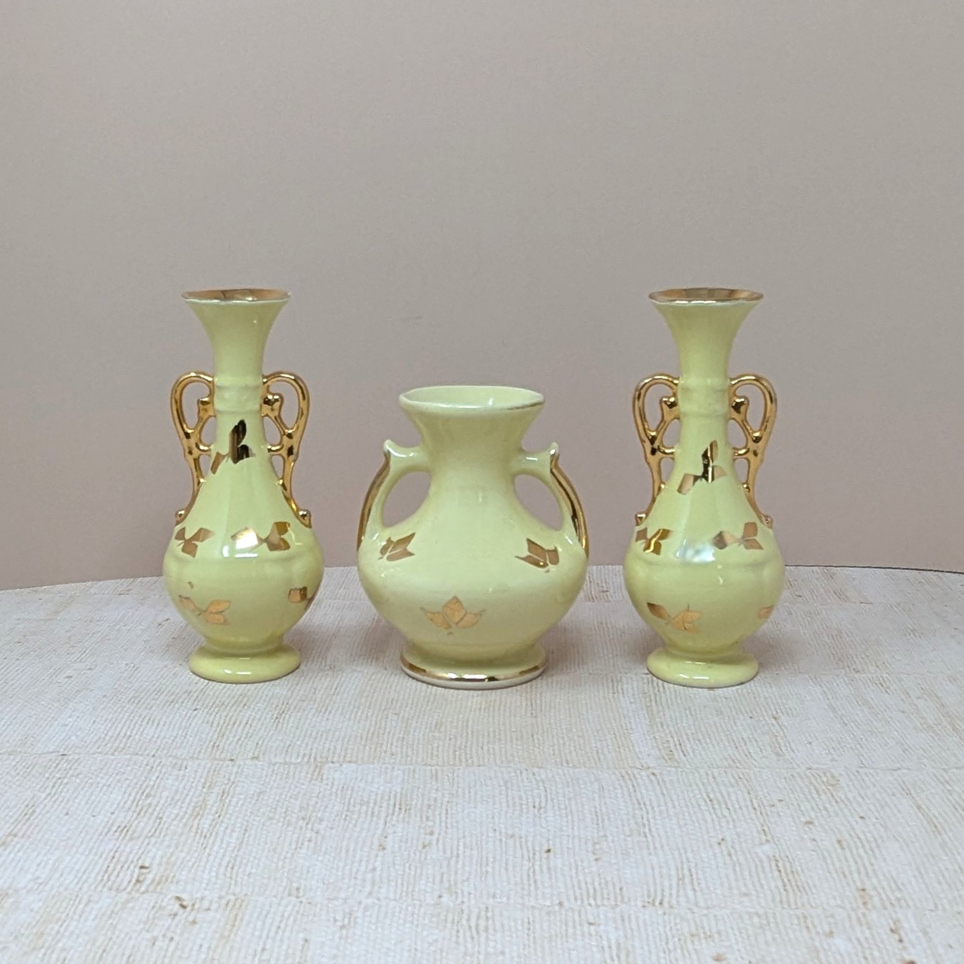 A collection of four vintage yellow ceramic vessels with bright gold accents. Includes a matching trio of Art Deco style vases with handles and gold leaf motifs, and a single rounded pitcher-style bud vase featuring a raised gold rose design.