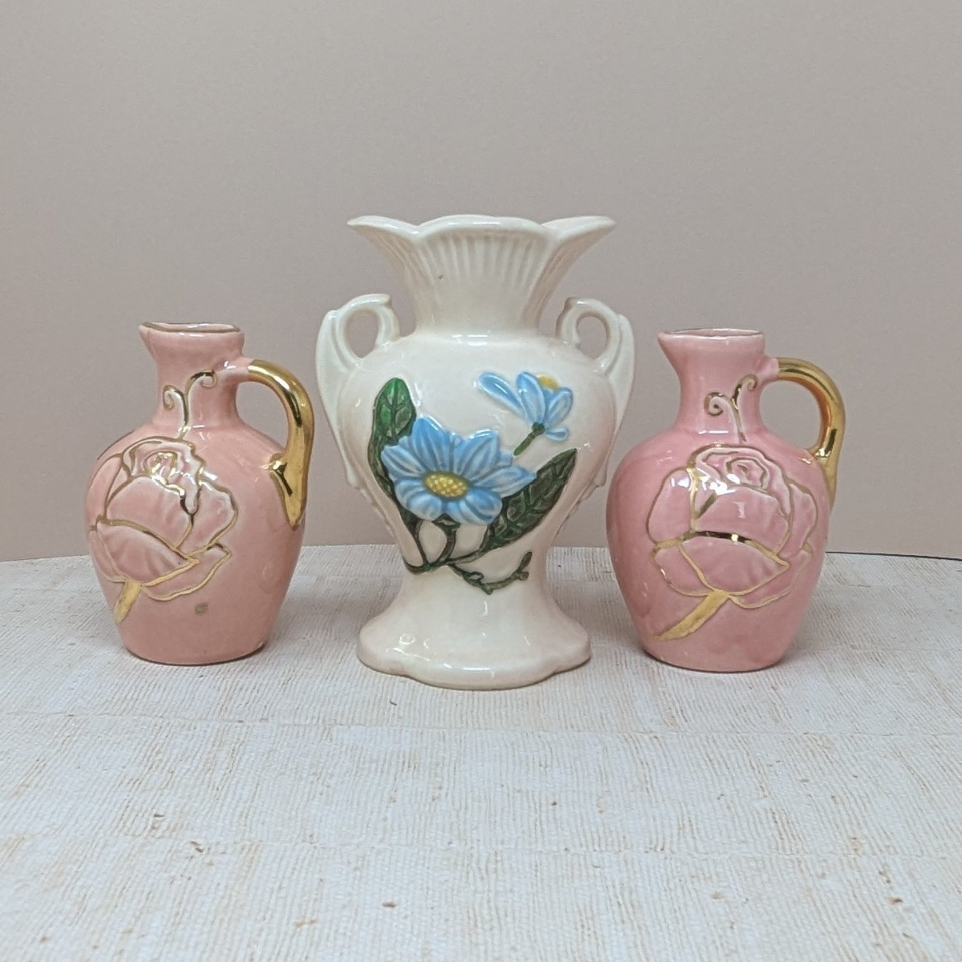 A three-piece vintage pink ceramic vase lot. The centerpiece is a 6.5-inch Hull Magnolia vase with blue flowers and double handles. It is flanked by a pair of 5-inch pink pitcher bud vases featuring embossed leaf designs and gold-clad handles.