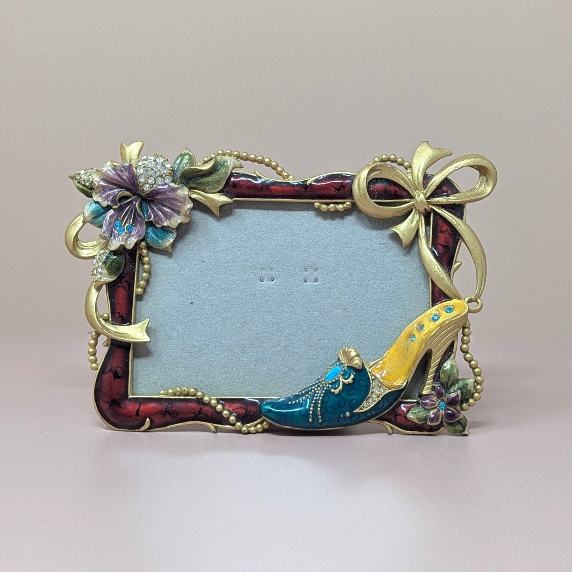 A horizontal photo frame with a deep red enamel border. Ornate gold-tone decorations include a crystal-studded purple hibiscus-style flower on the top left and a fashionable stiletto shoe on the bottom right.